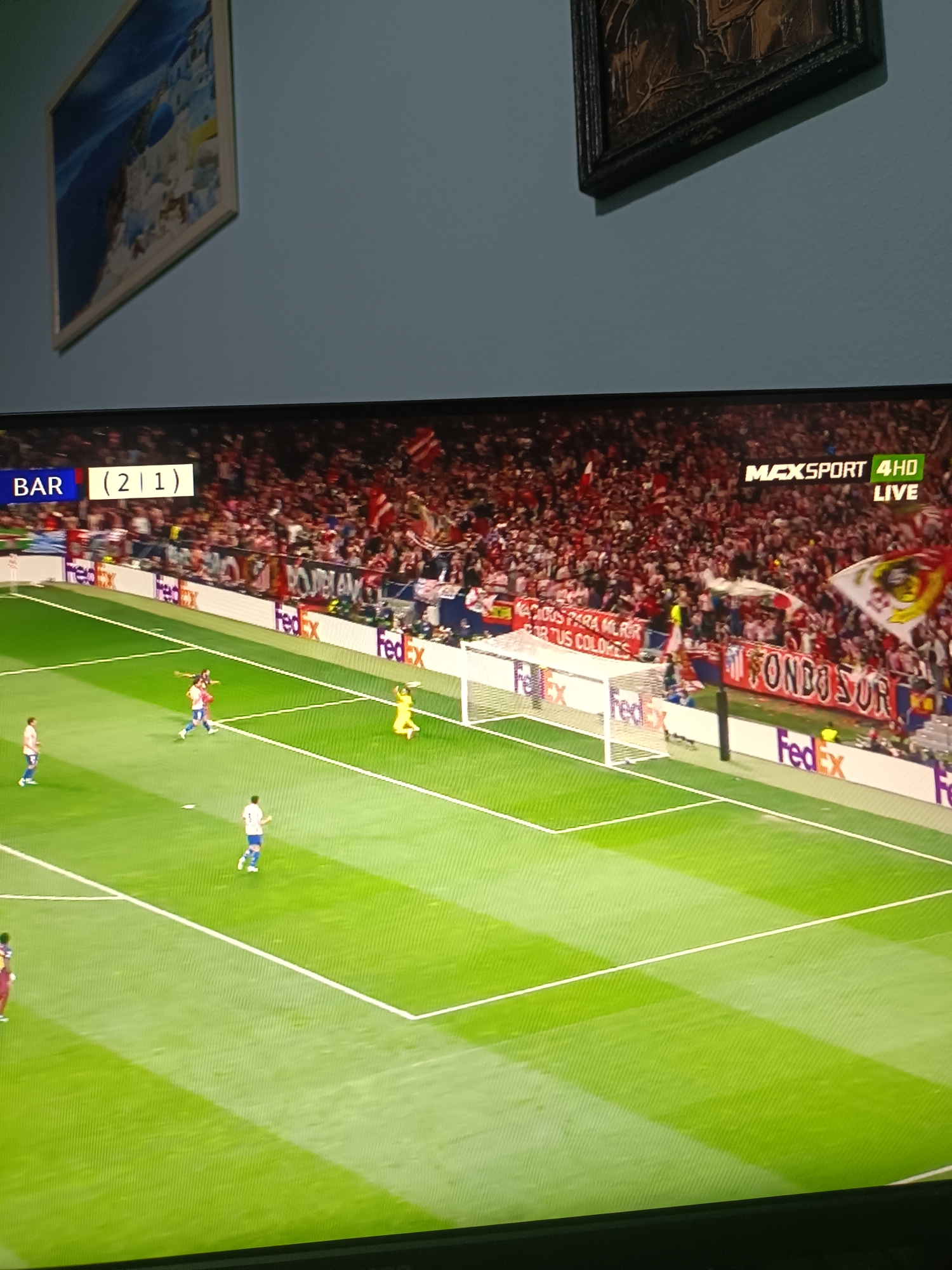 The image shows a television screen displaying a live soccer match. The score is BAR (2) - (1). The television is tuned to MAXSPORT 4HD. A crowd of spectators is visible in the background of the soccer field, with flags and banners displayed. Several players are on the field, with one near the goal being defended by the goalie. The advertising brand FedEx is displayed along the sidelines and behind the goalpost. There are two framed pictures hanging above the television. The location is not evident.