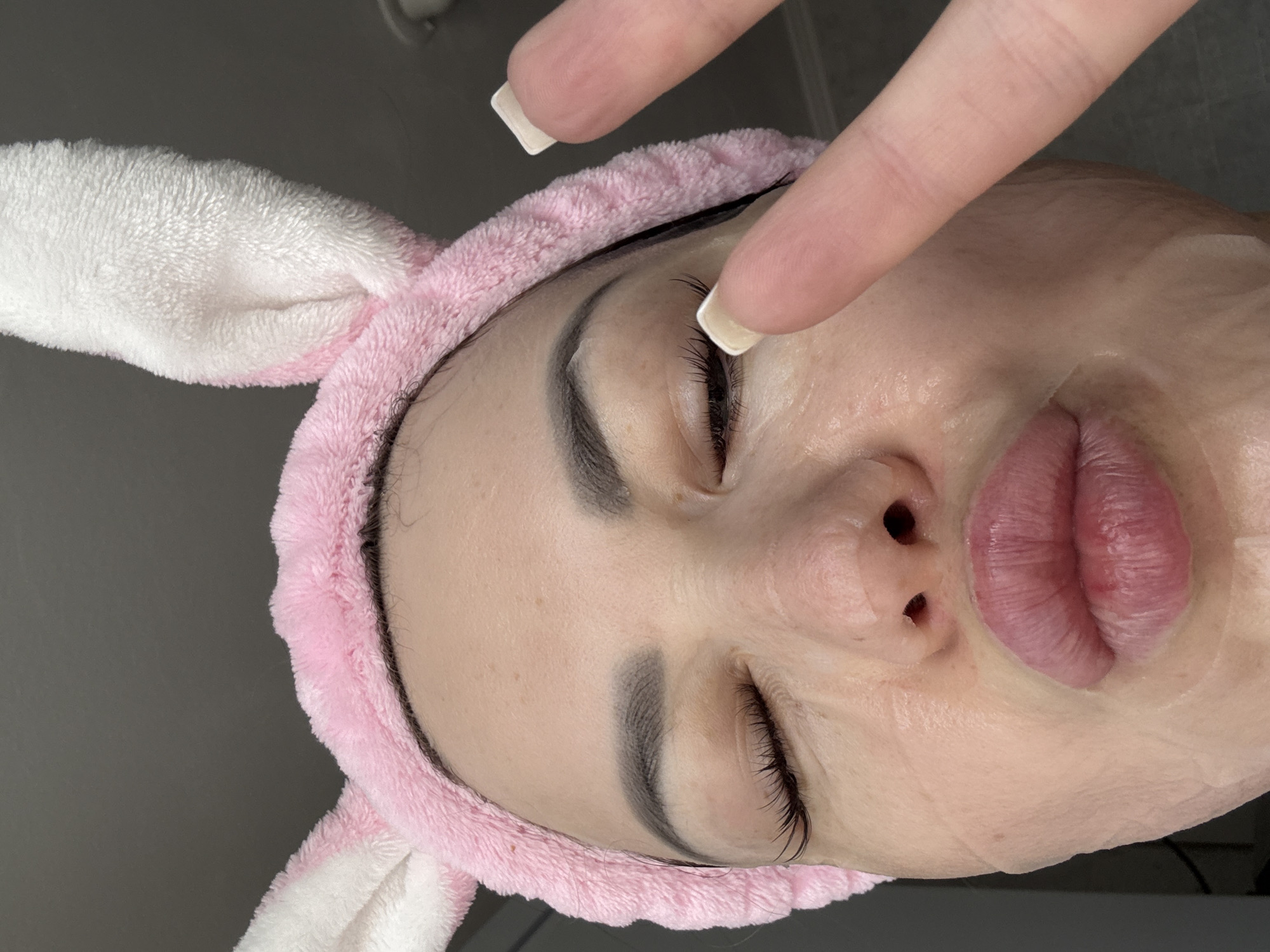 A close-up, downward-angled indoor shot depicts a person's face with their eyes closed, possibly resting or undergoing a beauty treatment. They are wearing a pink and white bunny ear headband. A semi-transparent sheet mask covers the majority of their face. The person's complexion appears light and somewhat shiny, possibly due to the mask. One hand, with polished square fingernails, is hovering near the top of the face. The background is plain and light gray.