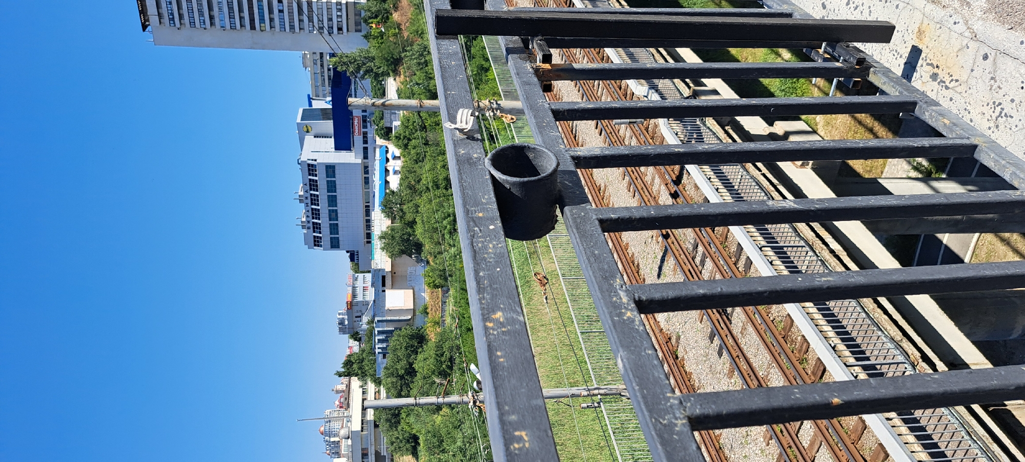 The image captures a clear, sunny day with a bright blue sky overlooking a cityscape. In the foreground, a black metal railing with visible rust spots runs across the bottom of the frame. A black pipe is attached to the top horizontal bar of the railing. Below the railing, a set of train tracks is visible, with ballast and sleepers laid out. Beyond the tracks, a lush green area with trees and bushes separates the tracks from the buildings in the background.
The city skyline features several buildings of varying heights and architectural styles. A tall, white high-rise building with numerous windows stands prominently on the right side. To its left, a multi-story building with a flat roof and rows of windows, possibly a commercial or office building, is visible, with the word "ПИРАМИДА" (Pyramida) written on its facade. Further to the left, other buildings and structures of different sizes and designs are scattered across the landscape, interspersed with more greenery. Utility poles and wires are also present in the scene, indicating urban infrastructure. The overall impression is one of an urban environment under a clear sky, with signs of both industrial activity (train tracks) and residential or commercial development.