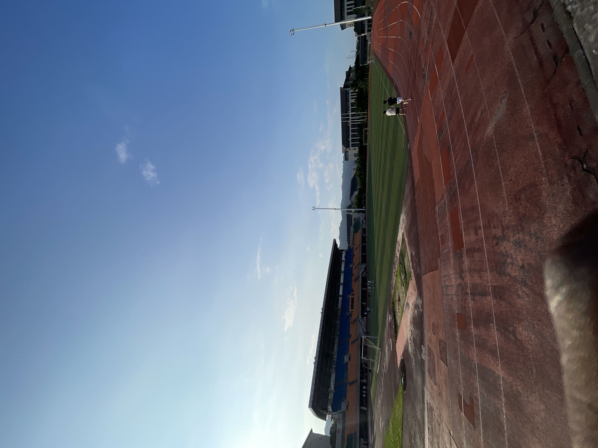 A vertically-oriented outdoor photograph depicts a stadium in Jiangjin, China. The sky is a clear blue with a few small clouds. On the right is a red running track. In the center is a green field with a soccer goal. Two people are visible on the track. A stadium with blue and orange seats is on the left. A row of buildings is in the background. Mountains are visible in the distance. Light poles are present. A wheel is partially visible in the lower right corner.