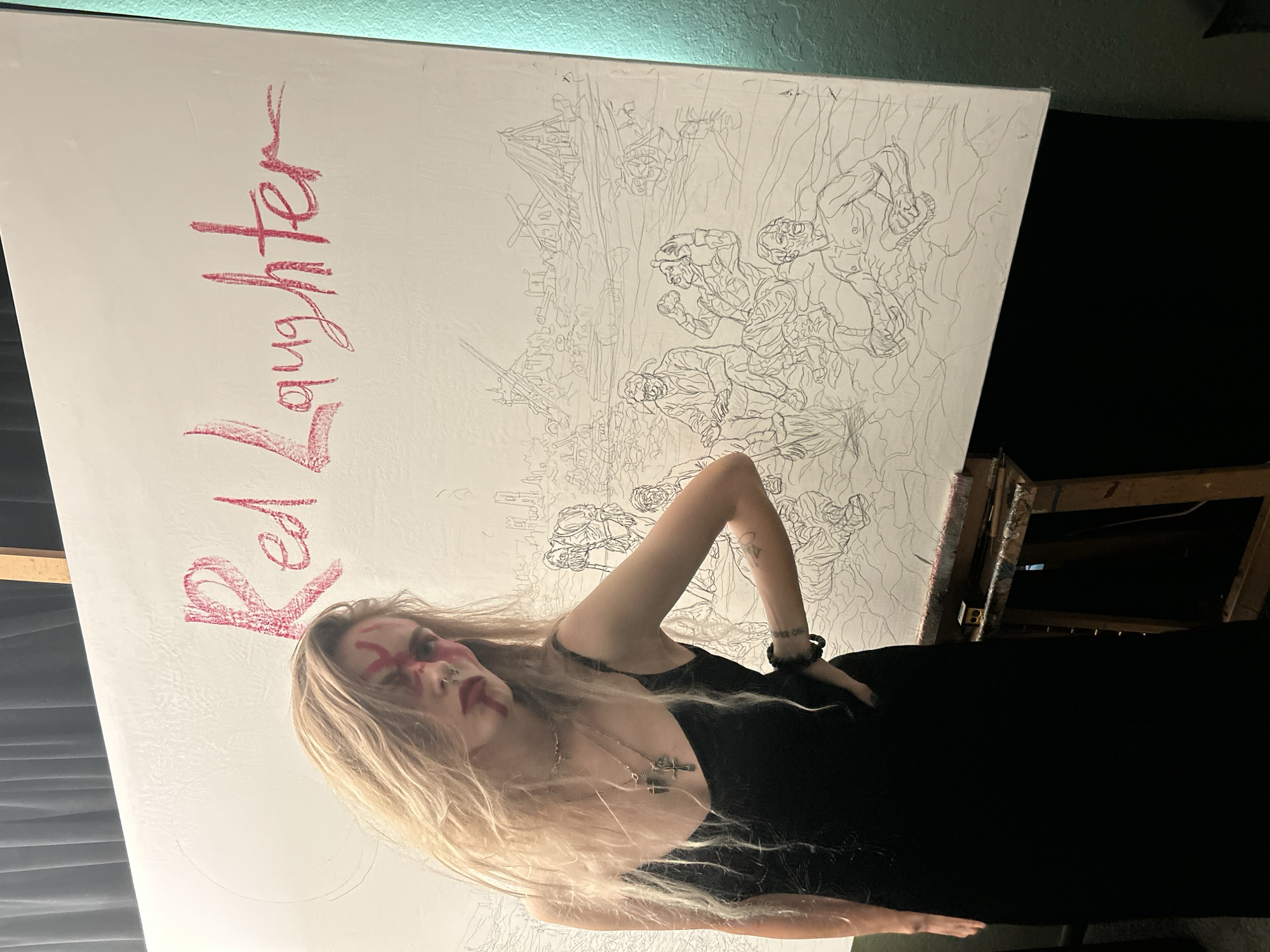 A low-angle indoor shot presents a young woman standing beside a large canvas. The canvas, which occupies a significant portion of the frame, displays a detailed line drawing, likely a war scene, alongside the words "Red Laughter" written in red. The woman has long blonde hair and is dressed in a black dress. She has red face paint. The setting appears to be an artist's studio or similar space, with a black curtain or backdrop visible behind and to the right of the canvas. The presence of the easel suggests a workspace dedicated to artistic creation. The image does not provide any information about the location in the city of Cape Coral, United States of America.