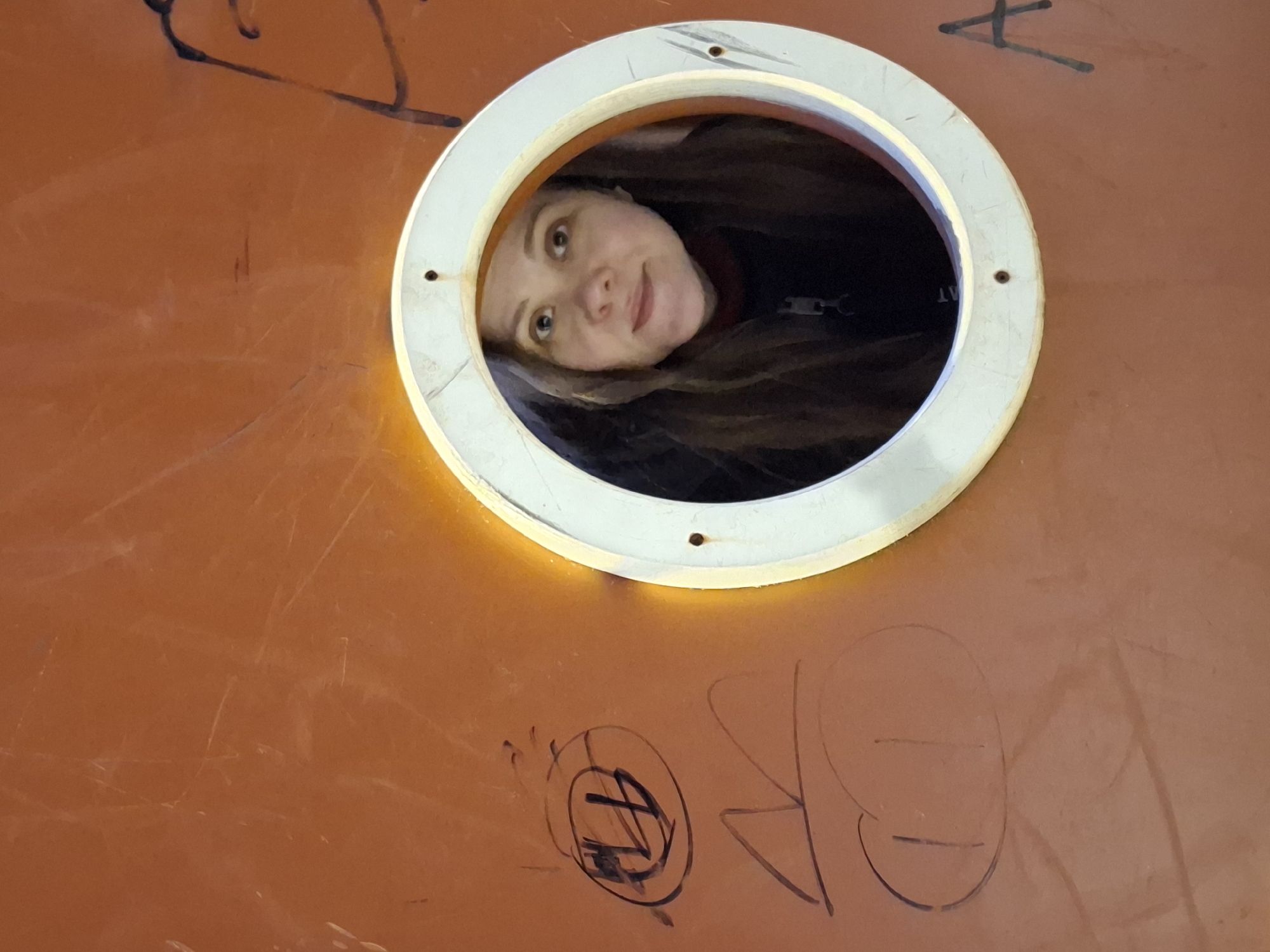 A woman with light skin and long brown hair peers through a circular opening, possibly a porthole or a viewing window, in an orange surface. The opening has a white frame secured with visible screws. Graffiti-like markings are present on the orange surface both above and below the opening. The location is Chaikovsky, Russia.