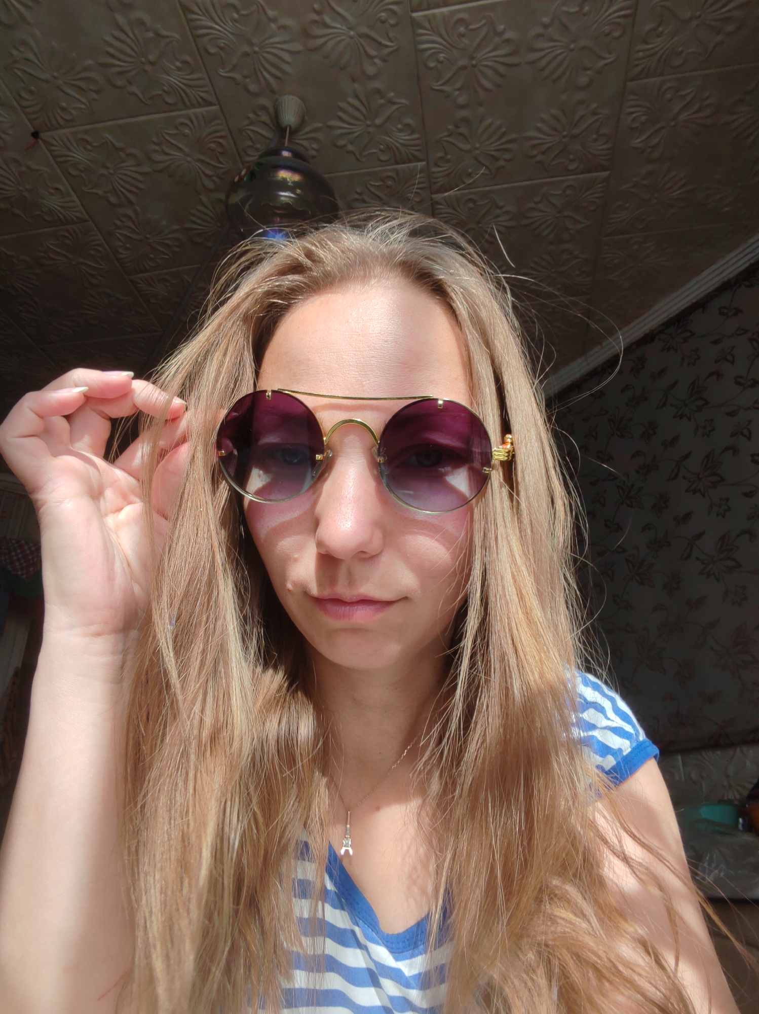 A blonde woman, wearing round, purple-tinted sunglasses and a blue and white striped shirt, is shown in a close-up, head-and-shoulders shot. She is holding her hair with her left hand. The background consists of a textured ceiling with floral patterns and a dark wall with a similar floral wallpaper. Sunlight casts strong shadows across the scene, particularly on the ceiling and the woman's face. A ceiling fan is visible in the upper left portion of the image. There is a small pendant in the shape of the Eiffel Tower on a necklace around her neck. The scene appears to be indoors, likely in a residential setting, and there are no visible indicators that place the location specifically within the city of Чухлэм, Russia.