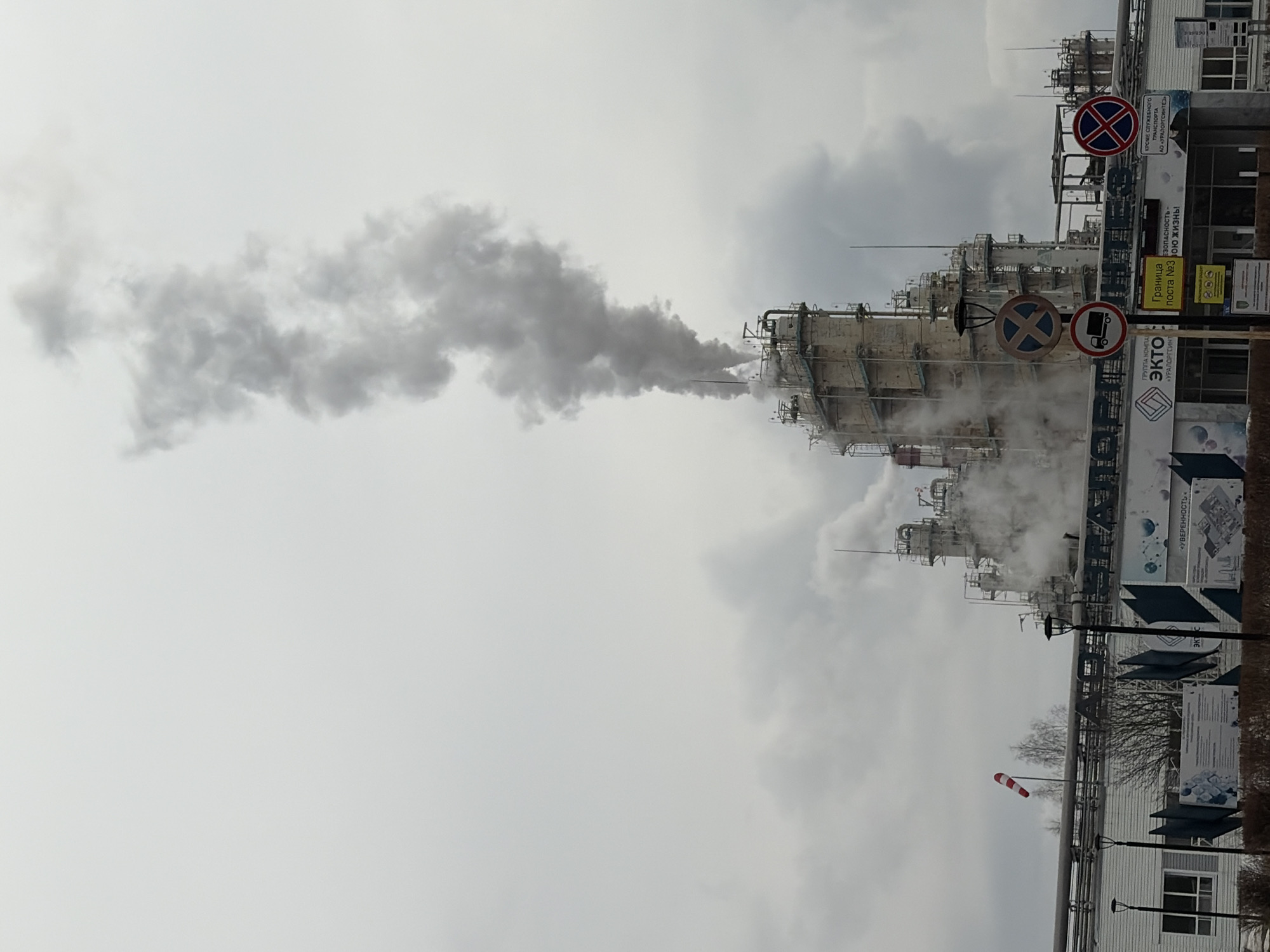 The image depicts an industrial scene, likely a chemical or petrochemical plant, located in Chernushka, Russia. A large, complex structure of pipes, tanks, and towers dominates the scene, with a plume of gray smoke or steam rising from one of the towers and dissipating into the overcast sky. Signage is visible, including warnings related to safety and restrictions on transportation. The name "EKTO" is prominent on a banner, potentially identifying the plant or its owner. The presence of a windsock indicates the potential for air quality concerns related to the emissions. The sky is overcast.