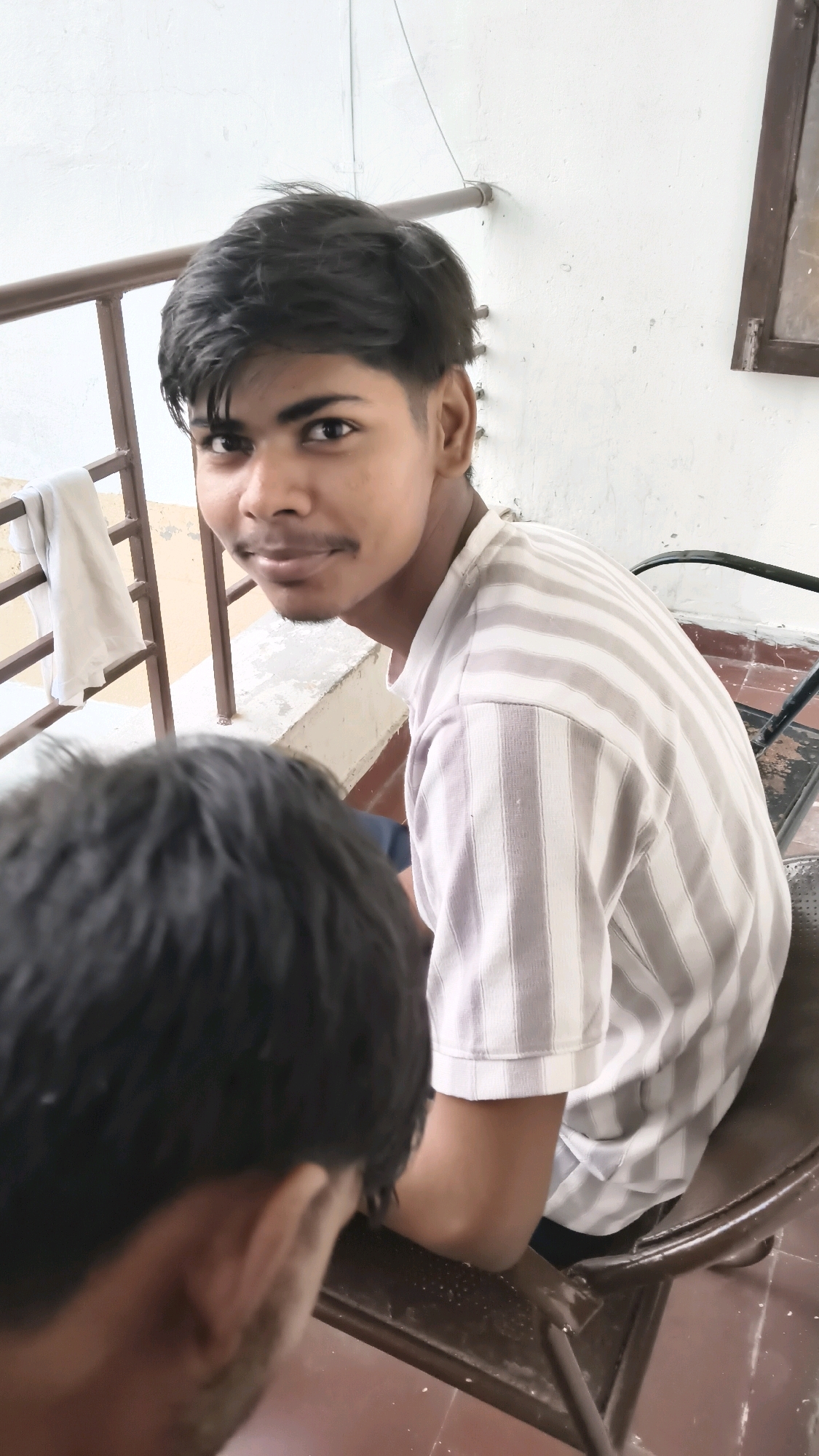 An individual, likely a young man, is seated in a chair facing towards the right of the frame, with his head turned to look back at the camera. He has dark, styled hair and is wearing a striped t-shirt. In the foreground, the back of another person's head and shoulders is visible, partially obscuring the seated individual. The setting appears to be outdoors, with a railing and a white wall in the background. A white cloth is draped over the railing. The overall environment suggests a casual or public space. The image does not provide specific visual cues to determine a location within the city of Libra, India.