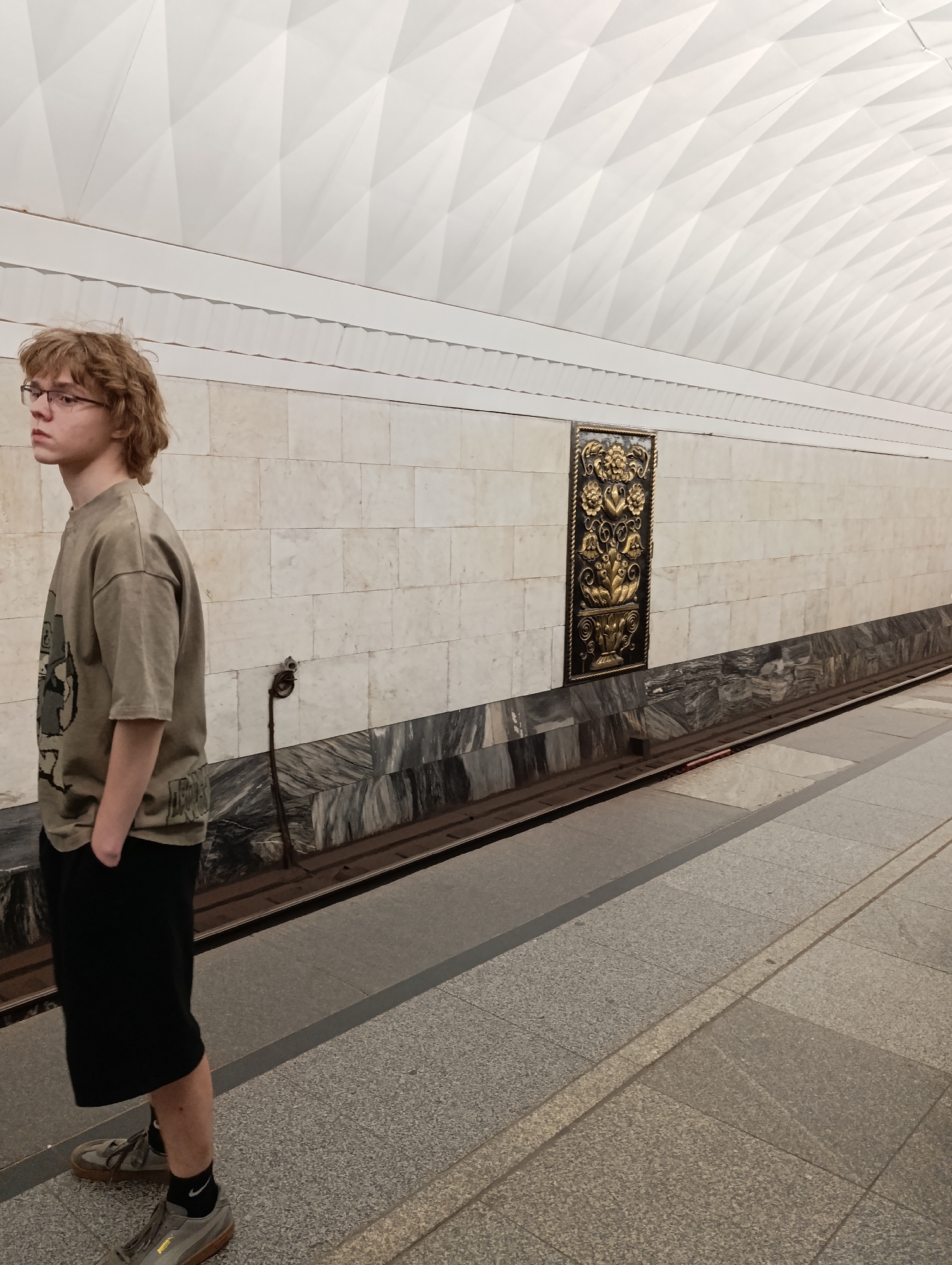 An indoor, eye-level image depicts a young male standing on a subway station platform in an unknown city. The male, positioned on the left side of the frame, has light brown, wavy, medium-length hair and wears dark-framed glasses. He is dressed in an olive green, loose-fitting, short-sleeved t-shirt with a faint graphic design on the front, black shorts, dark socks with a white Nike swoosh visible on the right ankle, and grey sneakers with brown soles. His hands are in his pockets, and his head is turned to the left, looking out of frame.
The platform wall behind him features two distinct sections: an upper part constructed of rectangular light beige tiles and a lower part of dark grey, marbled stone. A dark metal utility pole or lever-like device is affixed to the beige wall section, extending towards the tracks. Further down the platform, a rectangular ornate dark plaque with a prominent gold floral and foliate relief design is mounted on the beige wall. The platform floor is composed of grey square tiles, with a textured, light-colored tactile paving strip running parallel to the tracks. Two metal train tracks are visible alongside the platform. The ceiling is high, vaulted, and covered with white, diamond-patterned panels, creating a geometric texture. The scene is well-lit by internal lighting.