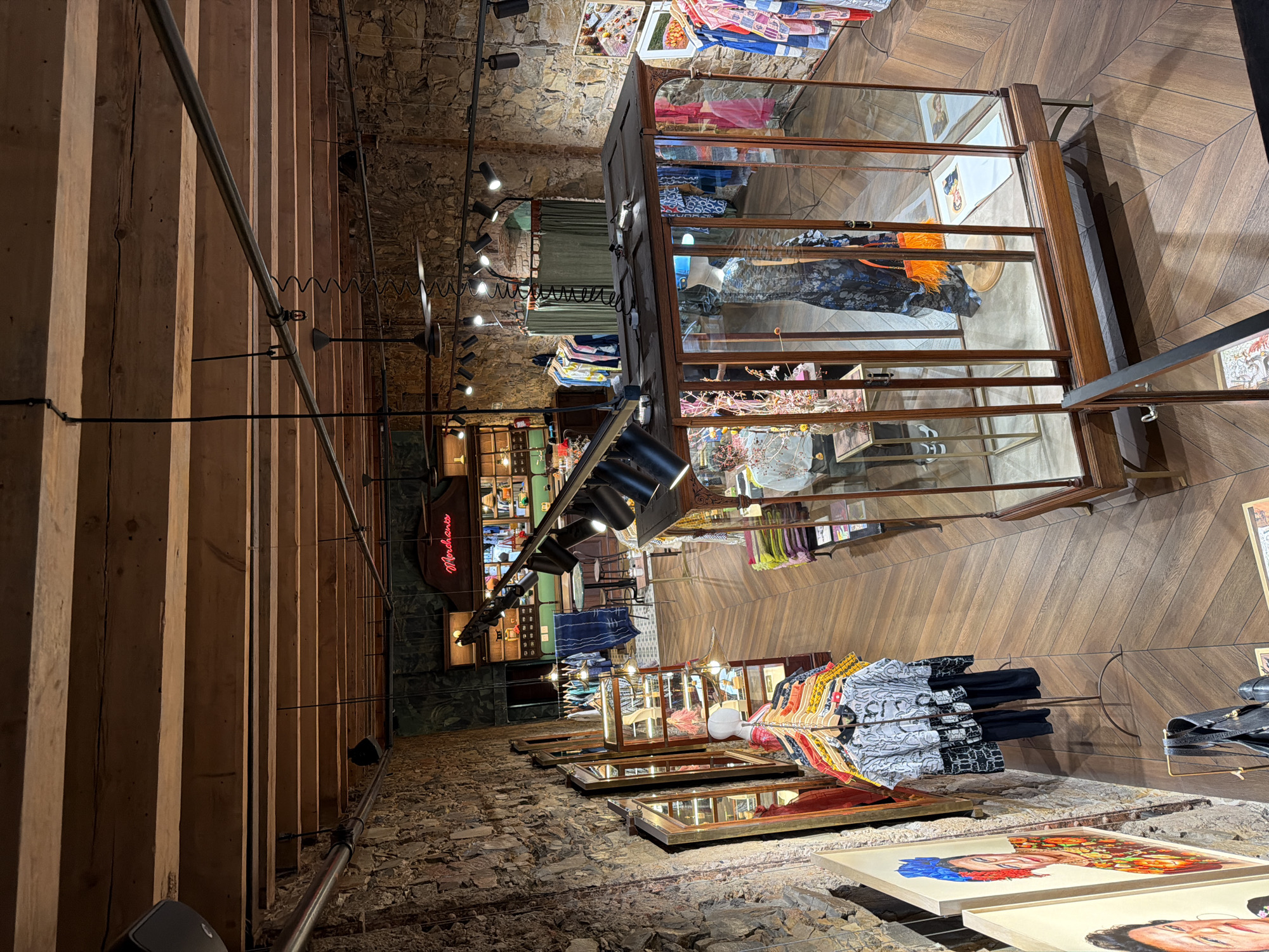 The image shows the interior of a retail store. It appears to be a clothing or design shop. The architecture of the store has exposed wooden beams and stone walls, suggesting a historic building. Clothing is displayed on racks and in glass display cases. There are also framed artworks on the floor near the foreground. A neon sign is present in the store. The location is Anthropologie & Co at the V&A Waterfront in Cape Town, South Africa.