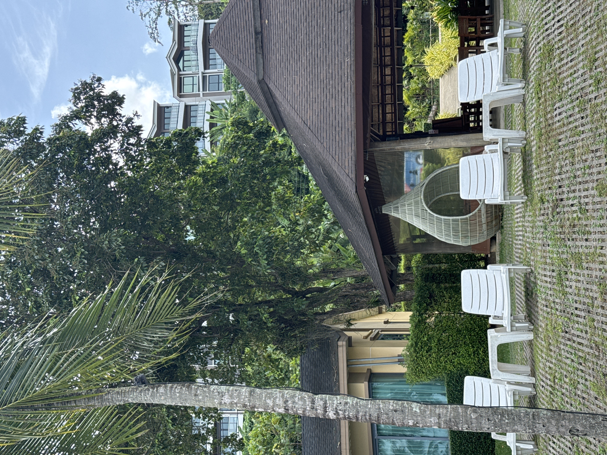 The image depicts an outdoor seating area with four white plastic lounge chairs and two small tables. In the background, a large, dark brown hexagonal roofed structure is visible, possibly a pavilion or restaurant. Beyond that, lush green trees and vegetation cover a hillside. Several multi-story buildings with balconies and modern architecture are situated higher up on the hill, suggesting a resort or residential complex. A tall palm tree trunk is prominent in the foreground on the left side of the image. The ground is covered with a patterned concrete or paver surface interspersed with patches of grass. The sky is visible with some clouds. The scene appears to be located in Pa Khlok, Thailand, likely at a resort given the style of buildings and the presence of lounge furniture. There are no people visible in the image.