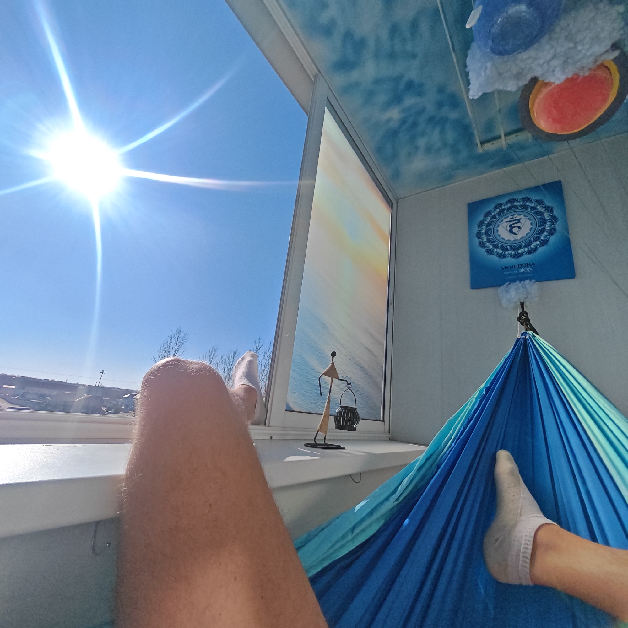 The image shows a person relaxing in a blue hammock inside a room. The person is wearing white socks, and their legs are visible, one extended towards the window. The room has a window with a view of a clear blue sky and a distant landscape with buildings, consistent with an urban setting. A small decorative sculpture sits on the windowsill next to a small black lantern. A blue poster with a circular design hangs on the wall above the hammock, next to a circular object with blue and red details on the ceiling. The ceiling is painted with a blue and white design resembling clouds. The location is specified as Rostov, Russia.