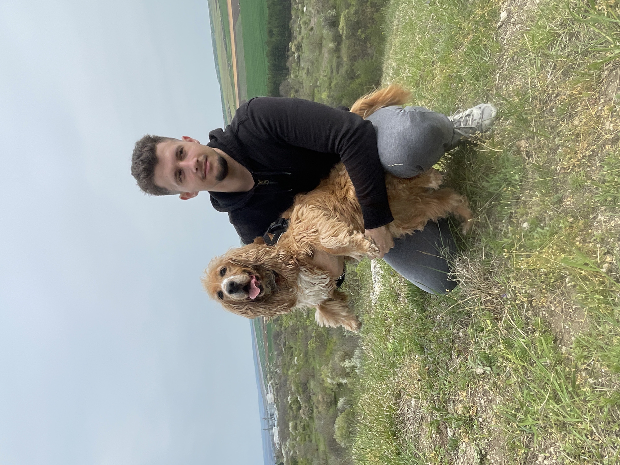 A man and a dog are outdoors in a grassy area. The man is light-skinned with short dark hair and a dark beard. He is wearing a black hoodie and gray jeans. He is squatting or kneeling and holding a medium-sized dog. The dog is a golden color, likely a Cocker Spaniel, with its tongue sticking out. They are on a hill or higher elevation as a landscape with fields and trees can be seen in the background. The sky is overcast. The location is possibly in the Razgrad area of Bulgaria. The environment is grassy.