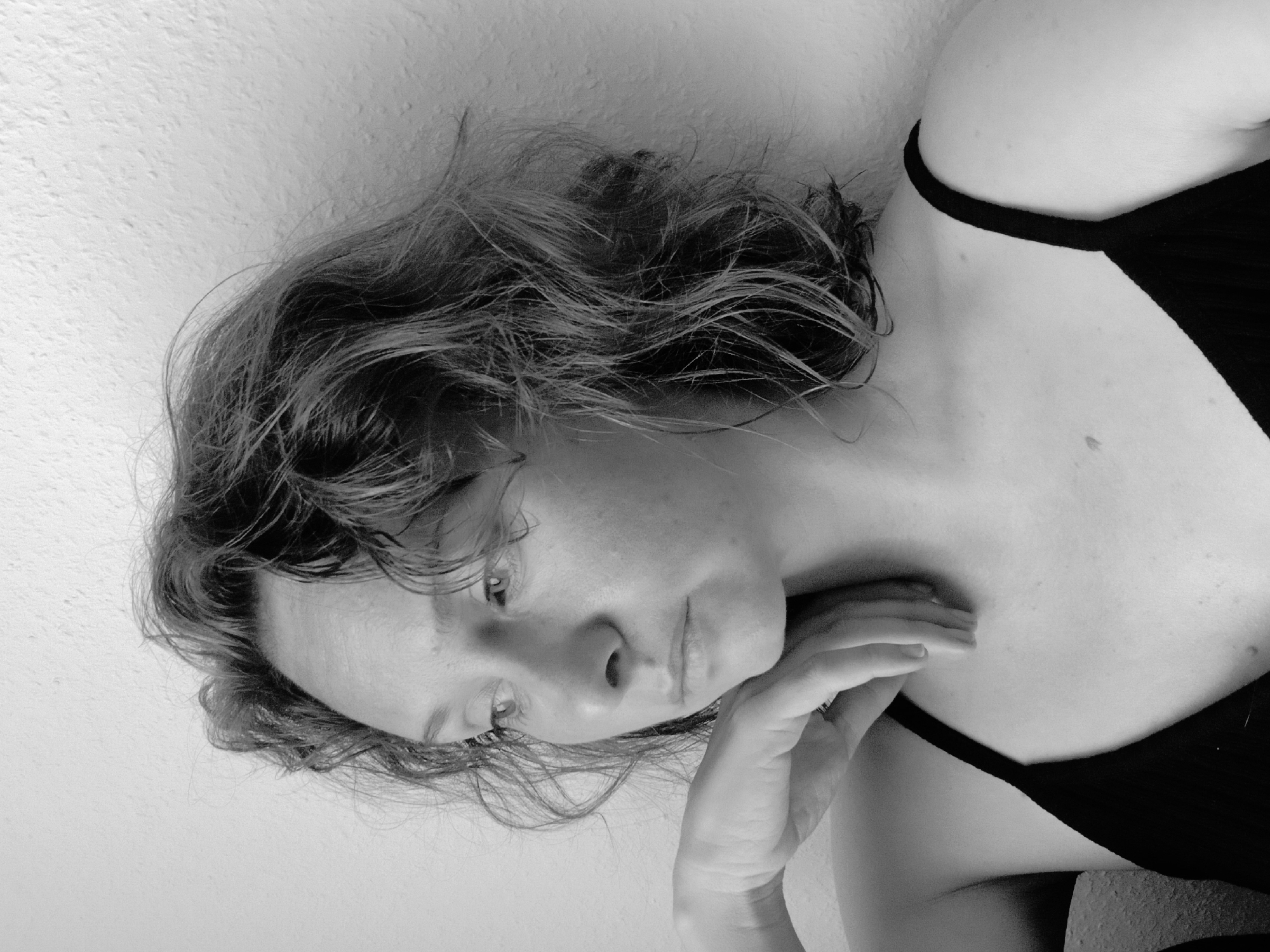 A black and white close-up portrait of a young Caucasian woman is shown. She has curly, shoulder-length brown hair and is looking to her left. Her left hand is resting on her chin and neck. She is wearing a black tank top. The background is a textured, light-colored wall. The image is cropped to show the woman from the chest up. The location of this image is not determinable from the visual information provided.
