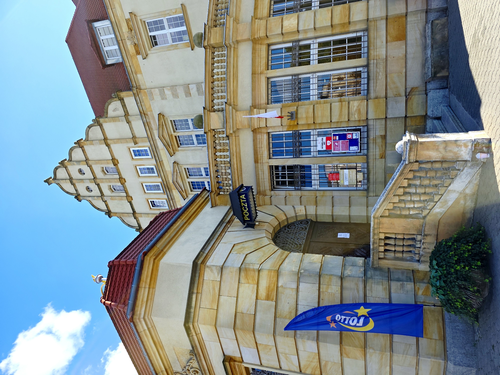 The image shows the facade of the main post office building in Poznan, Poland, located on Plac Wolności. The building exhibits a historical architectural style, characterized by its sandstone cladding, ornate detailing, and a red tiled roof. In front of the building, a blue flag with a yellow logo featuring stars and the word "TOLLO" stands to the left. A set of stone stairs with a decorative balustrade leads up to the main entrance, which features a large wooden door with intricate metalwork. Above the entrance, a sign with the word "POCZTA" indicates it is a post office. The facade also includes numerous large windows, some of which have white frames and decorative metal grates. A Polish flag is mounted on the wall to the right of the entrance. The sky is bright blue with a few white clouds. The ground in front of the building is paved with interlocking bricks, and a bush is planted in a large pot to the left of the stairs. Several posters are affixed to the windows. The lighting suggests it is daytime with bright sunlight casting shadows.