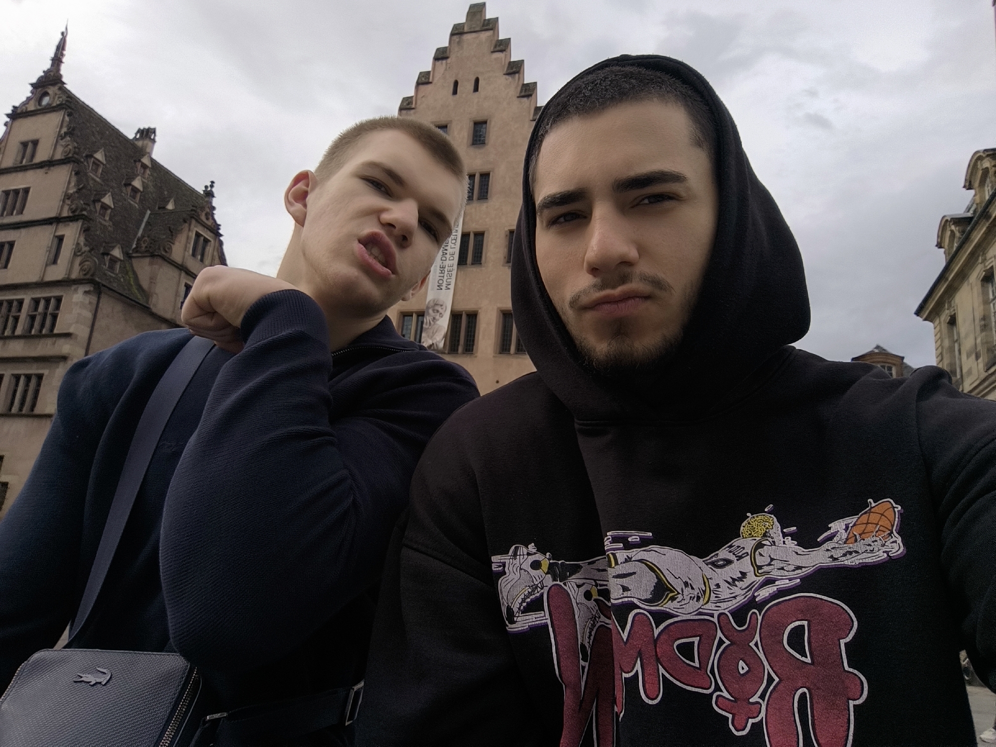 Two young men pose for a selfie in Strasbourg, France, with the historical Maison Kammerzell building visible behind them. The man on the right wears a black hoodie with a graphic design. The man on the left wears a dark blue sweater and has a black Lacoste cross-body bag. They are outside in overcast weather.