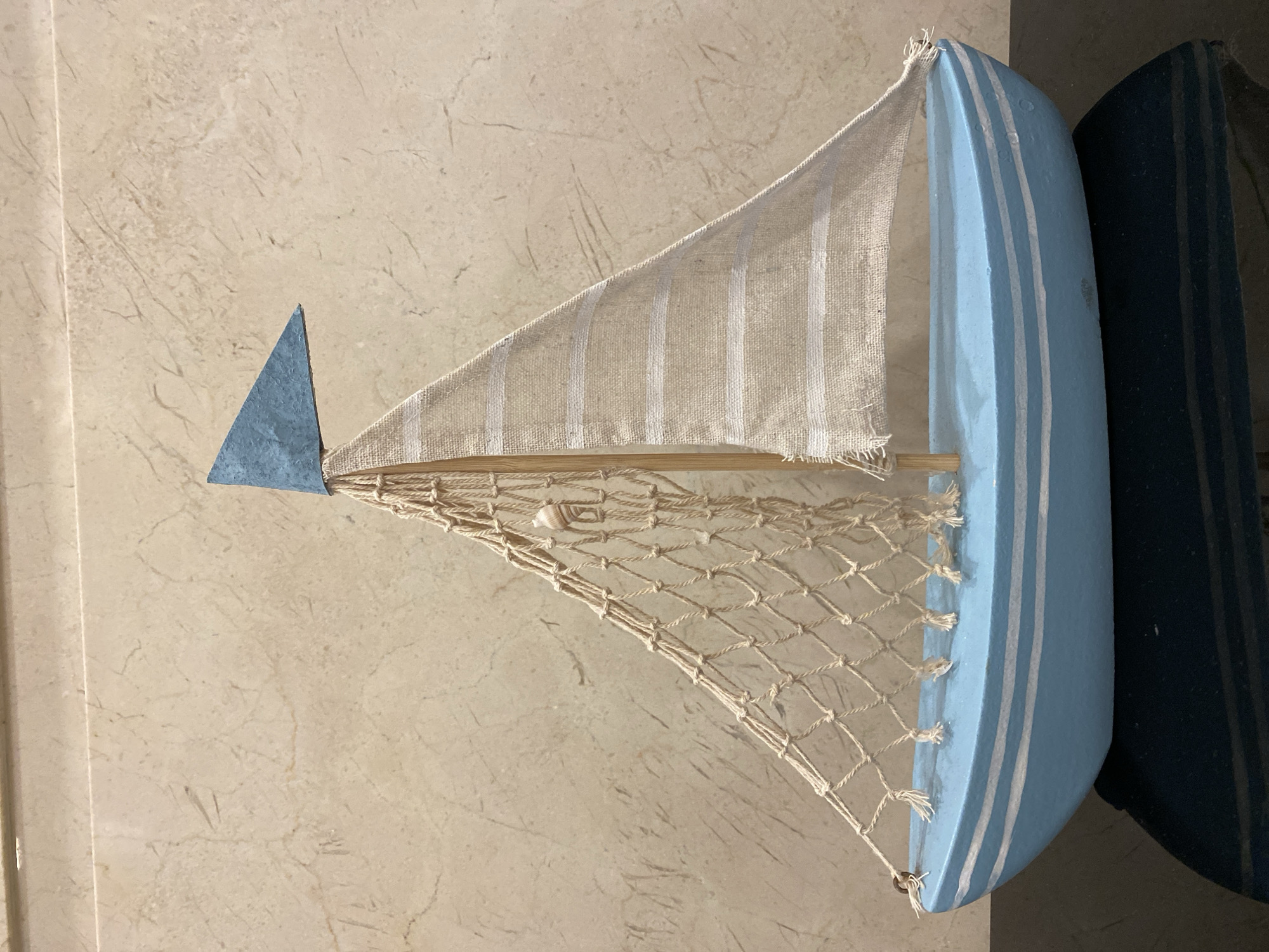 A small, light blue wooden sailboat model with white stripes is displayed against a textured, beige wall. The sailboat has a cream-colored net attached to its mast, which also holds a small, light blue triangular flag. A tiny seashell is caught in the net. The boat sits on a dark, reflective surface. The scene is indoors and does not appear to be located in Da Nang, Vietnam.