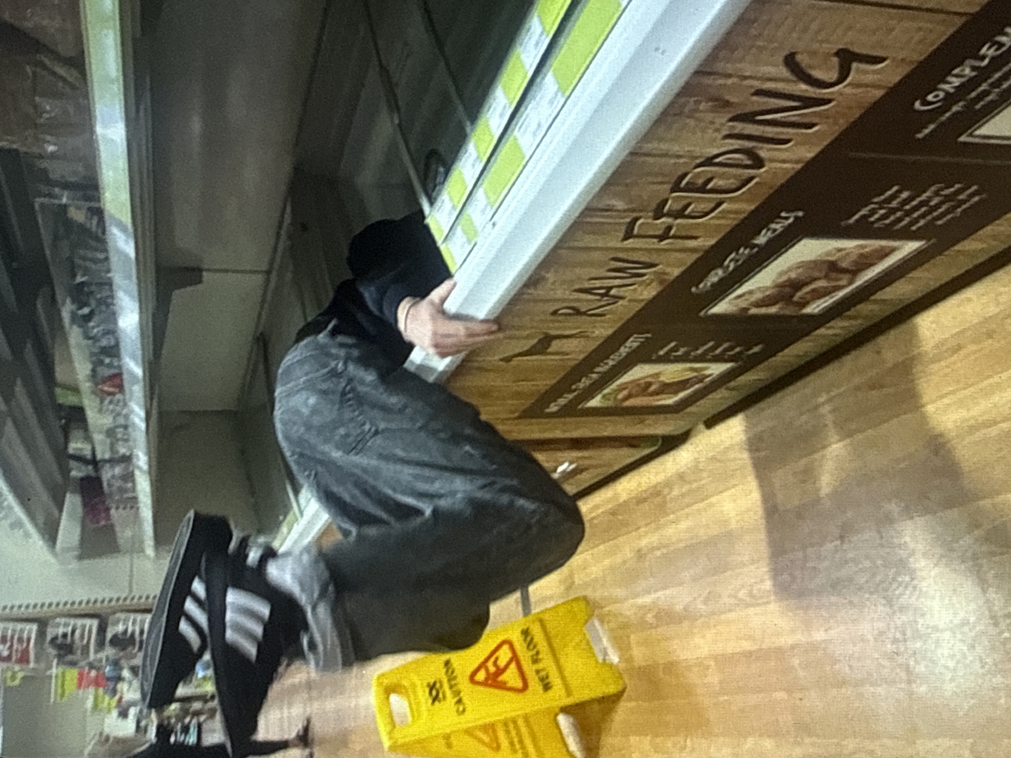 An individual is depicted partially inverted, with their upper body submerged into an open freezer chest in a retail setting in the city of Kardzhali, Bulgaria. The freezer's visible exterior features a wooden-texture decal with "RAW FEEDING" text and images of pet food products, indicating a pet supply section. The individual's legs, clad in dark pants and black Adidas slide sandals with white stripes, extend upwards from the freezer. Their right hand is visible gripping the top edge of the freezer. A yellow A-frame "CAUTION WET FLOOR" sign is positioned on the light-colored wooden floor to the left of the freezer. In the background, another person's lower legs and feet, wearing light-colored sandals, are visible on the left side of the frame, walking away. Overhead shelves, containing various indistinct packaged items, are visible in the background.