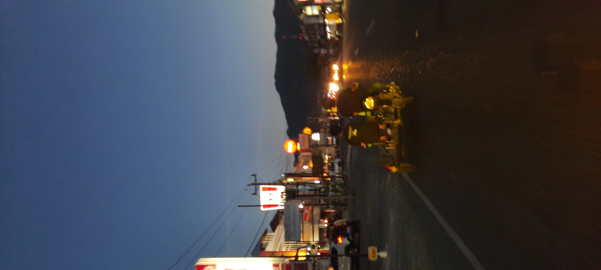 The image depicts a street scene at dusk in Lambheu, Indonesia. A three-wheeled vehicle, likely a motorcycle with a sidecar, is being ridden down the road. Two individuals are visible on the vehicle, one driving and another seated behind. The vehicle appears to be carrying some equipment or goods in the sidecar.
On the left side of the road, there are buildings with illuminated signs, including what appears to be a KFC restaurant and other businesses. Power lines are visible overhead. In the background, a mountain range is silhouetted against the darkening sky. The road surface is asphalt and shows signs of being wet, possibly from recent rain. Streetlights and the headlights of other vehicles are contributing to the illumination of the scene. The overall atmosphere is that of a busy commercial area as night begins to fall.