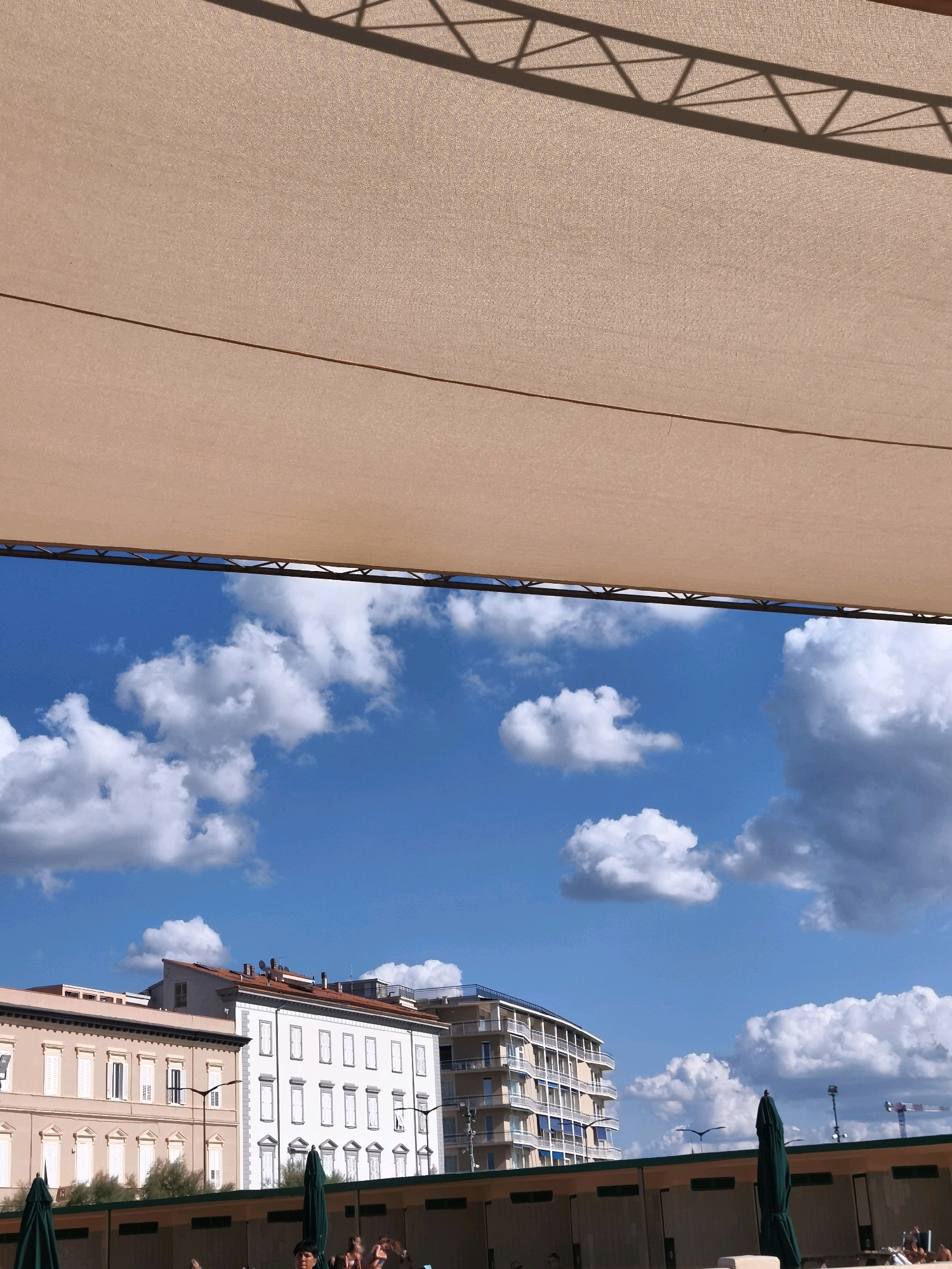 This image, taken in Livorno, Italy, depicts a sunny day with a bright blue sky filled with scattered white clouds. In the foreground, a large, light-colored awning stretches across the top of the frame, casting a geometric shadow from its supporting structure onto its surface. Below the awning, buildings with multiple windows and balconies are visible. To the left, a pale pink building with white shutters lines the street. Adjacent to it is a white building with a more ornate facade and similar shutters. Further to the right, a modern multi-story building with balconies and blue awnings stands out. At the bottom of the frame, a row of structures, possibly changing rooms or cabanas, are visible, with dark green umbrellas in front of them. A few people are present, some partially visible in the lower right corner, appearing to be relaxing or sunbathing. The scene suggests a coastal or beachside location within the city.