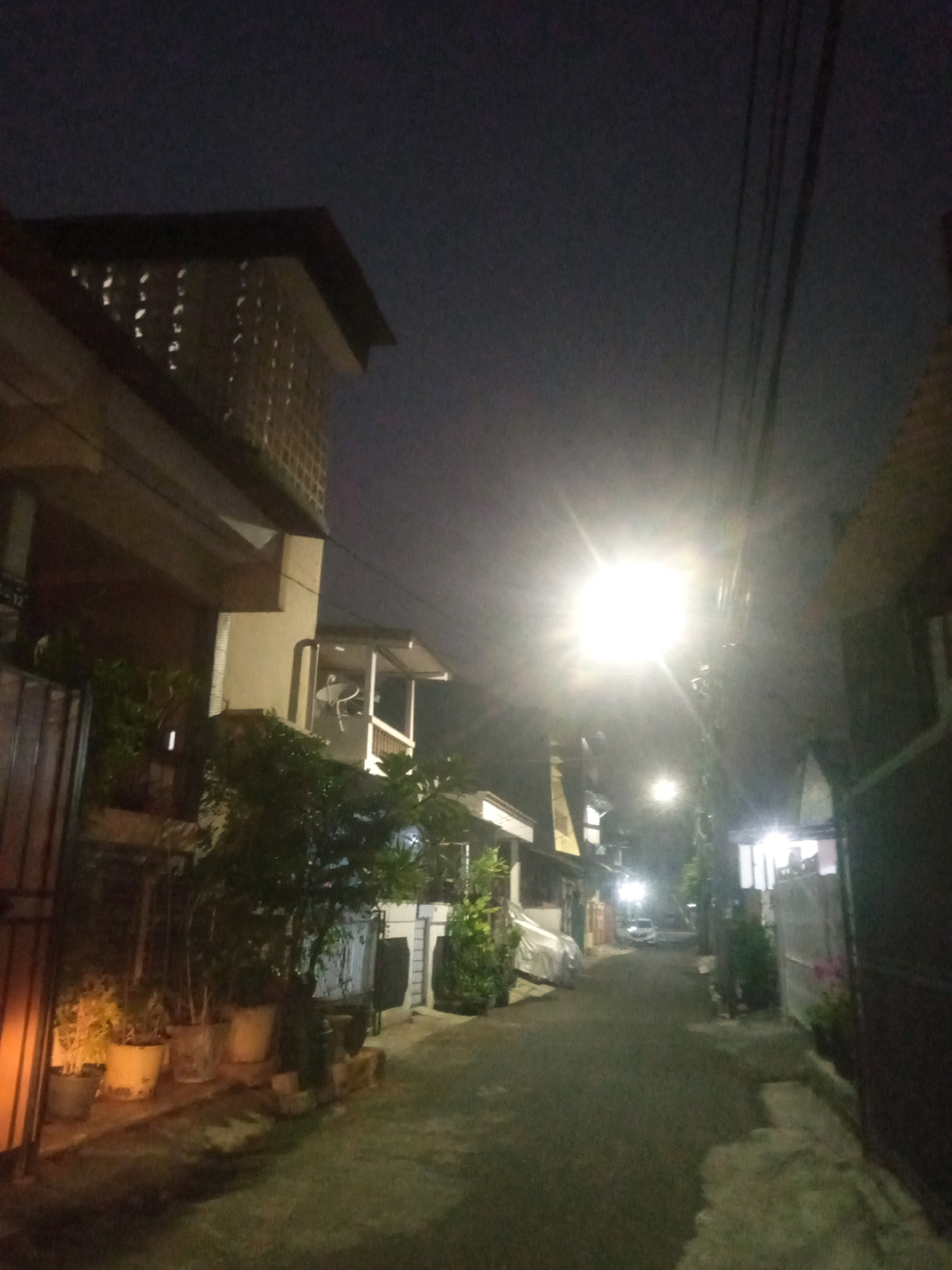 The image depicts a dimly lit residential alleyway in North Jakarta, Indonesia, at night. A bright street light illuminates the scene, casting long shadows. On the left side of the alley, a building with several windows and a slanted roof is visible. Potted plants line the walkway in front of the building. The alley itself is paved and curves into the distance, where another street light and the silhouette of a car can be seen. The right side of the alley is bordered by another building with a dark facade. Power lines are strung overhead. The overall atmosphere is quiet and still, typical of a residential area at night.