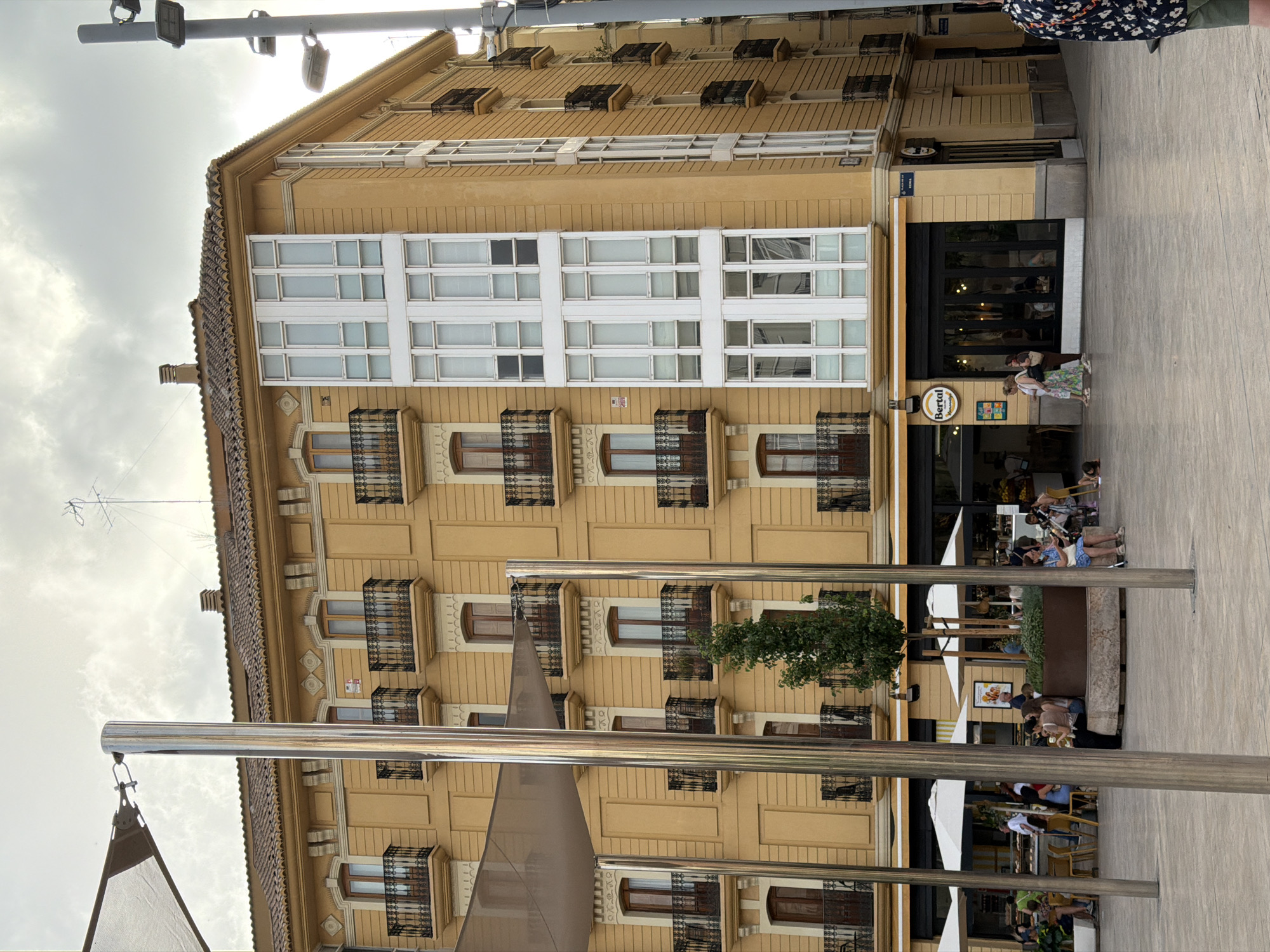 This is a vertical, eye-level, full-shot photograph of a yellow building on a cloudy day. The building has multiple floors with balconies and windows. The ground floor features a storefront with large glass doors and windows. In front of the building, there is a plaza with people sitting at tables under umbrellas and walking. There are also several metal poles and a shade structure visible. The overall environment appears to be an urban setting with a commercial area.