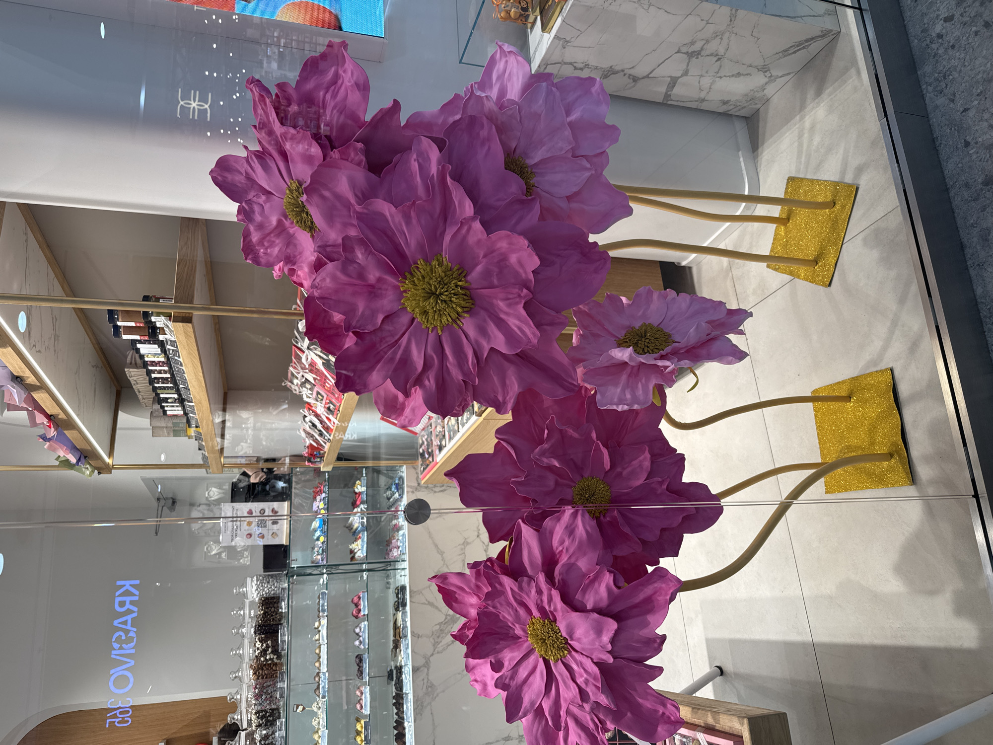 The image shows a storefront display of large, artificial pink flowers with yellow centers and glittery gold bases. The storefront is a confectionery or sweets shop with glass display cases filled with chocolates and candies. The name "Krasivo" is visible on the glass, written in Cyrillic. The location is likely in Saint Petersburg, Russia. The overall environment is bright and modern, typical of a retail setting.