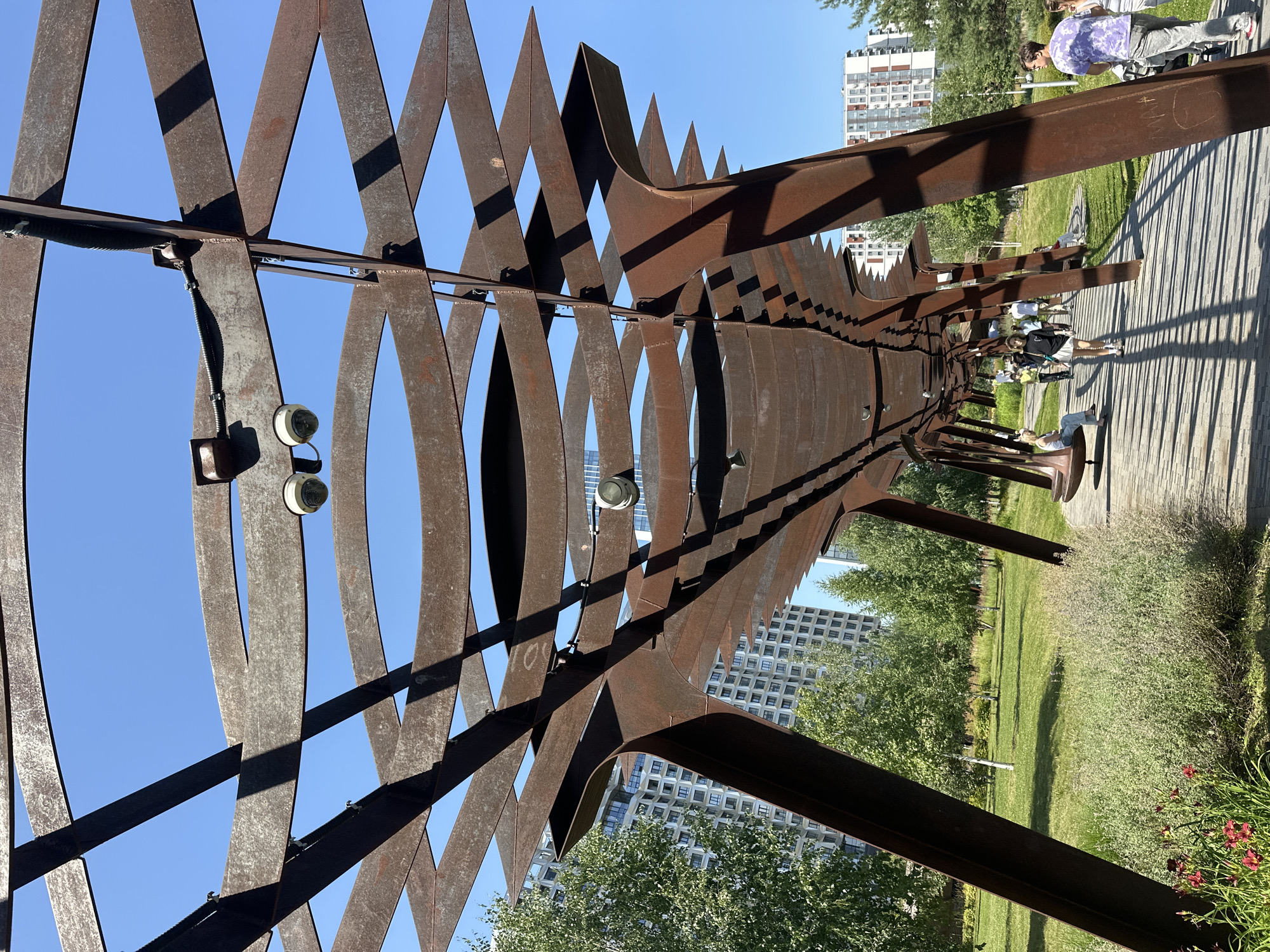 The image depicts a modern park with a distinctive rusty metal canopy overhead. The canopy is an intricate structure of curved and angled metal beams, creating a rhythmic pattern against the clear blue sky. Two security cameras are visible attached to the underside of the canopy. A paved walkway runs beneath the canopy, casting long shadows from the metalwork. Several people are walking along the path, some paused to rest or observe the surroundings. One person is seated on a curved bench attached to a canopy support. Beyond the walkway and canopy, a well-maintained grassy area is visible, dotted with trees and bordered by modern apartment buildings. In the foreground, lush green bushes and some red flowers add a touch of natural color to the scene. The architecture suggests a contemporary urban park setting. This appears to be a scene from the Zaryadye Park in Moscow, Russia, known for its modern design and integration of natural elements with urban infrastructure.