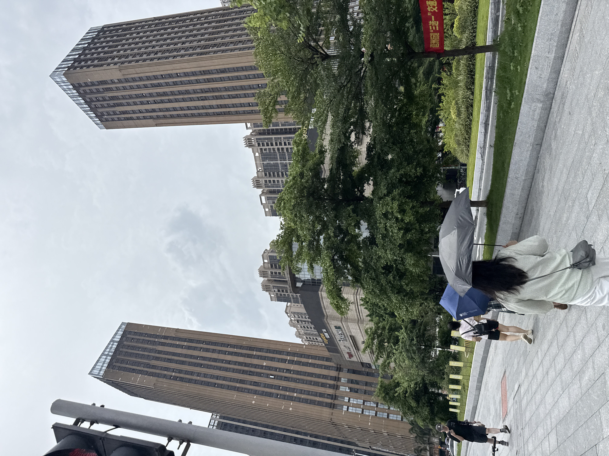 In Haizhu District, China, on a cloudy day, two tall, modern buildings dominate the skyline, their facades a pattern of beige panels and dark windows. In front of these structures, a multi-story building, possibly a shopping center, displays illuminated signage, including "T FRESH" and "getha". Lush green trees partially obscure the view of these buildings, their leaves forming a dense canopy. On a paved sidewalk in the foreground, four individuals walk away from the camera. One person, dressed in black shorts and a black t-shirt, walks ahead with a phone in hand and a device resembling a drone backpack. Behind them, two people hold umbrellas, one blue and one gray, as they walk with a third person in a light-colored top and white pants carrying a gray crossbody bag. The sidewalk is made of large, rectangular gray tiles, and a grassy median with landscaping separates the walkway from a road where a white vehicle is visible. A red banner with Chinese characters hangs on a tree to the right of the frame. A traffic light is visible on the left side of the image.