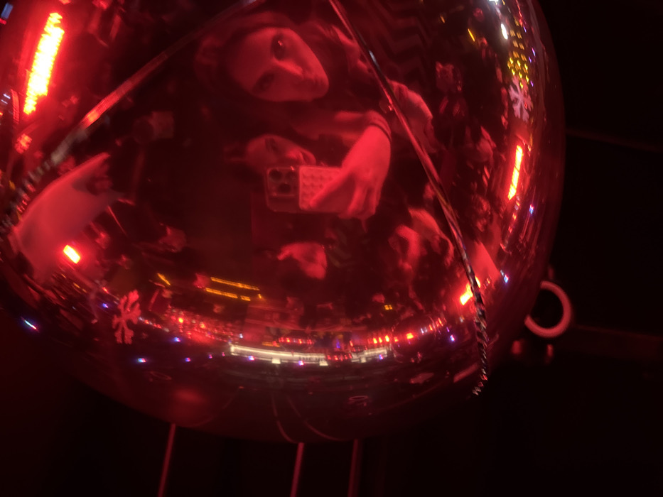 The scene is captured through the reflection on a large, spherical, highly polished or mirrored object, predominantly illuminated by intense red light. Within this distorted reflection, two individuals are clearly discernible. One person, positioned centrally, is holding a smartphone, actively photographing the reflection; their face is partially obscured by the device. To the right, the face of a second individual is distinctly visible, looking towards the reflective surface. Numerous light sources, appearing as streaks and points of red, blue, purple, and orange, are reflected across the sphere, with one reflection resembling a snowflake pattern. A metallic chain and a ring are visible at the bottom of the spherical object, indicating it is suspended or affixed. The environment appears dark, with the reflective surfaces capturing the scattered light. The scene is located in San Bernardino Tlaxcalancingo, Mexico.