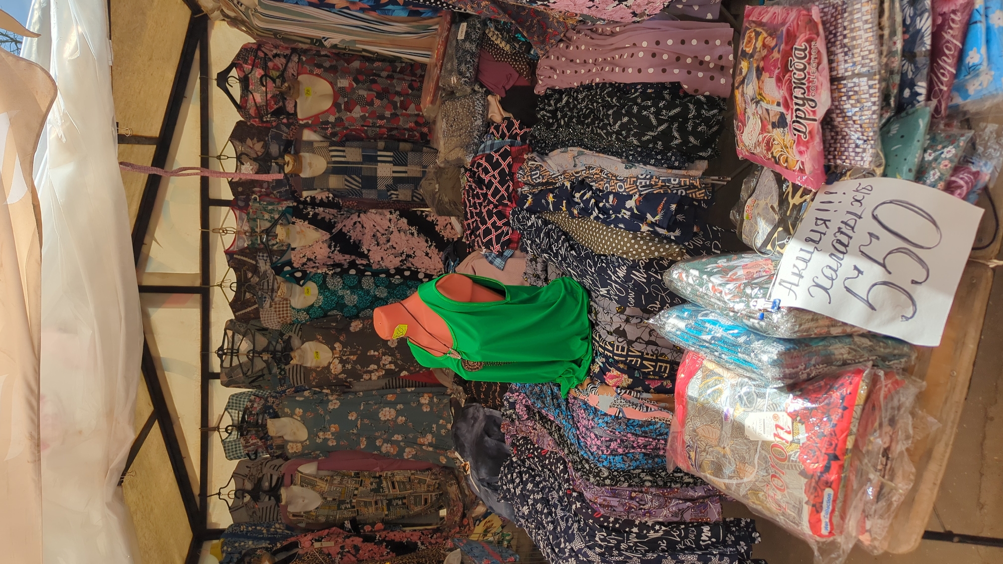 The image depicts an outdoor market stall in Kurovskoye, Russia, displaying a variety of clothing items. The stall is covered with a white tarp, supported by wooden beams. Racks of hanging dresses are visible at the top, featuring various floral and geometric patterns. Below, the merchandise is laid out on tables and the ground, with a significant emphasis on dresses and other garments in vibrant colors and diverse prints.
A mannequin dressed in a bright green sleeveless top with a necklace is positioned prominently in the center of the stall. In the foreground, several packaged clothing items, possibly robes or bathrobes, are stacked on a table. A handwritten sign with Russian text, "Акция!!! Халаты!!!" (Sale!!! Robes!!!), and the price "550" is displayed next to these packages. The overall scene suggests a bustling marketplace with an array of textiles and apparel for sale.