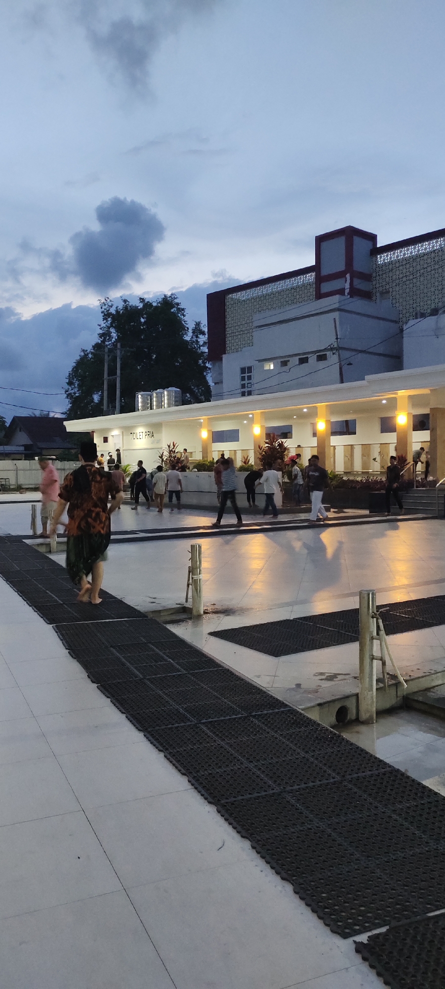 In the city of Bireuenmeunasahdayah, Indonesia, a group of people are gathered near a building with a sign that reads "TOILET PRIA". The scene appears to be at dusk or early evening, with a cloudy sky overhead. The building has a modern design with white walls, a red trim, and decorative grilles. Yellow lights illuminate the covered walkway in front of the building.
Several individuals are visible in the image, some walking, others standing or interacting. One person in the foreground is walking barefoot on a black, grated walkway. This walkway leads towards the building where other people are congregated. The ground is a light-colored tile. In the background, a large tree stands near the building, and houses are visible to the left. The overall atmosphere suggests a public space where people are either arriving, departing, or socializing.