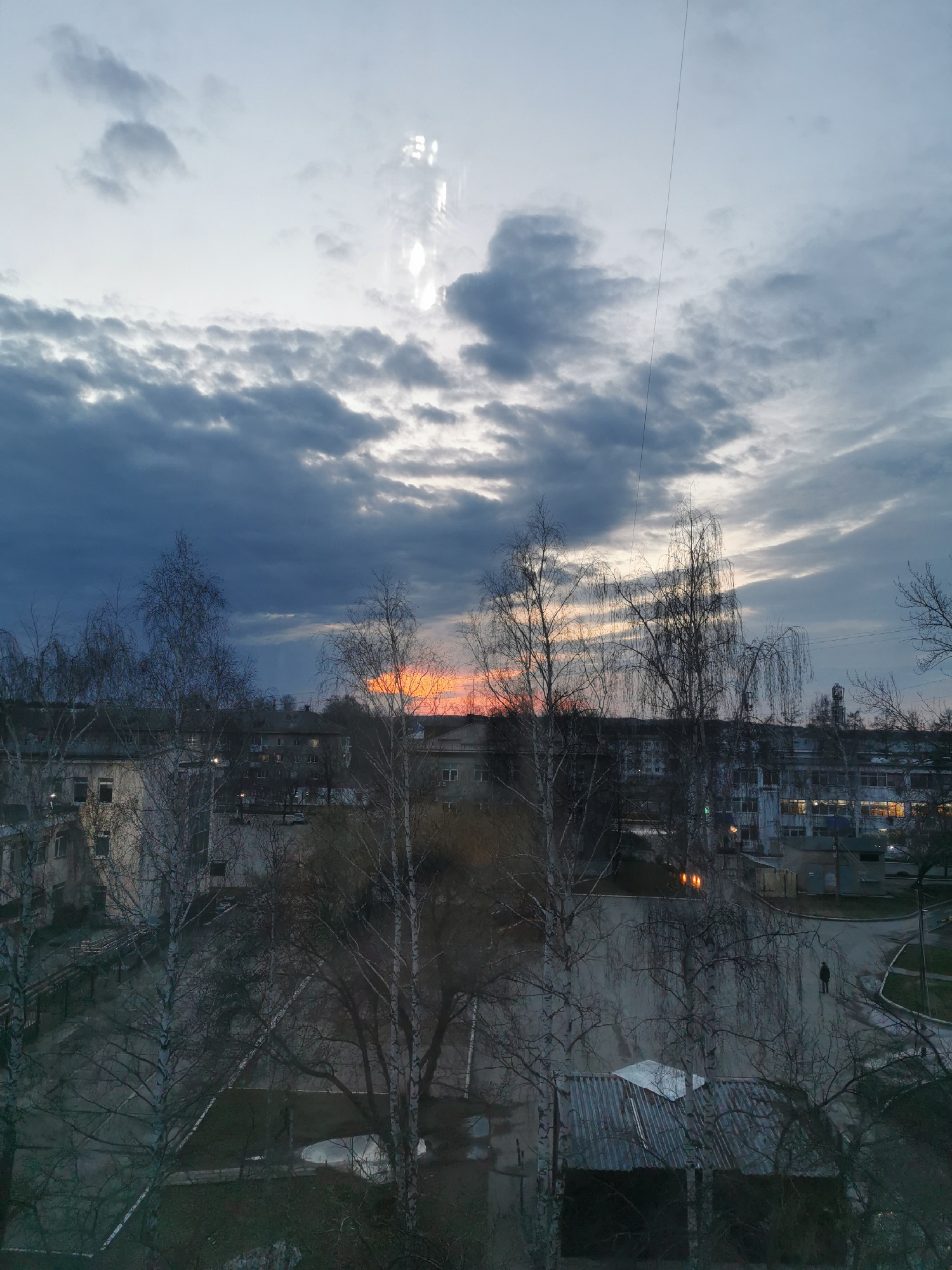 The image presents an overcast sky with a band of orange sunset light near the horizon, visible through a silhouetted treeline. The scene is set in a city neighborhood, indicated by the presence of multi-story buildings with illuminated windows in the background. In the foreground, bare trees without leaves frame the view of a gray, paved area. A single person is visible on the right side of the frame, walking on the pavement. A low-lying structure with a metal roof is also present in the lower part of the image.