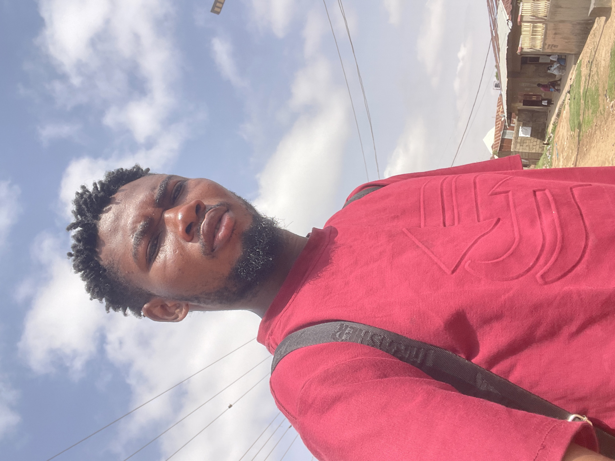 A medium-range shot looking up at a man in Unguwar Gabas, Nigeria. The man is Black, has short, curly hair, a beard, and is wearing a red t-shirt and a black strap over his shoulder. The sky is visible in the background with white clouds, along with power lines. In the background on the right is a building with a corrugated metal roof. Two people are seated in the building, one wearing red.