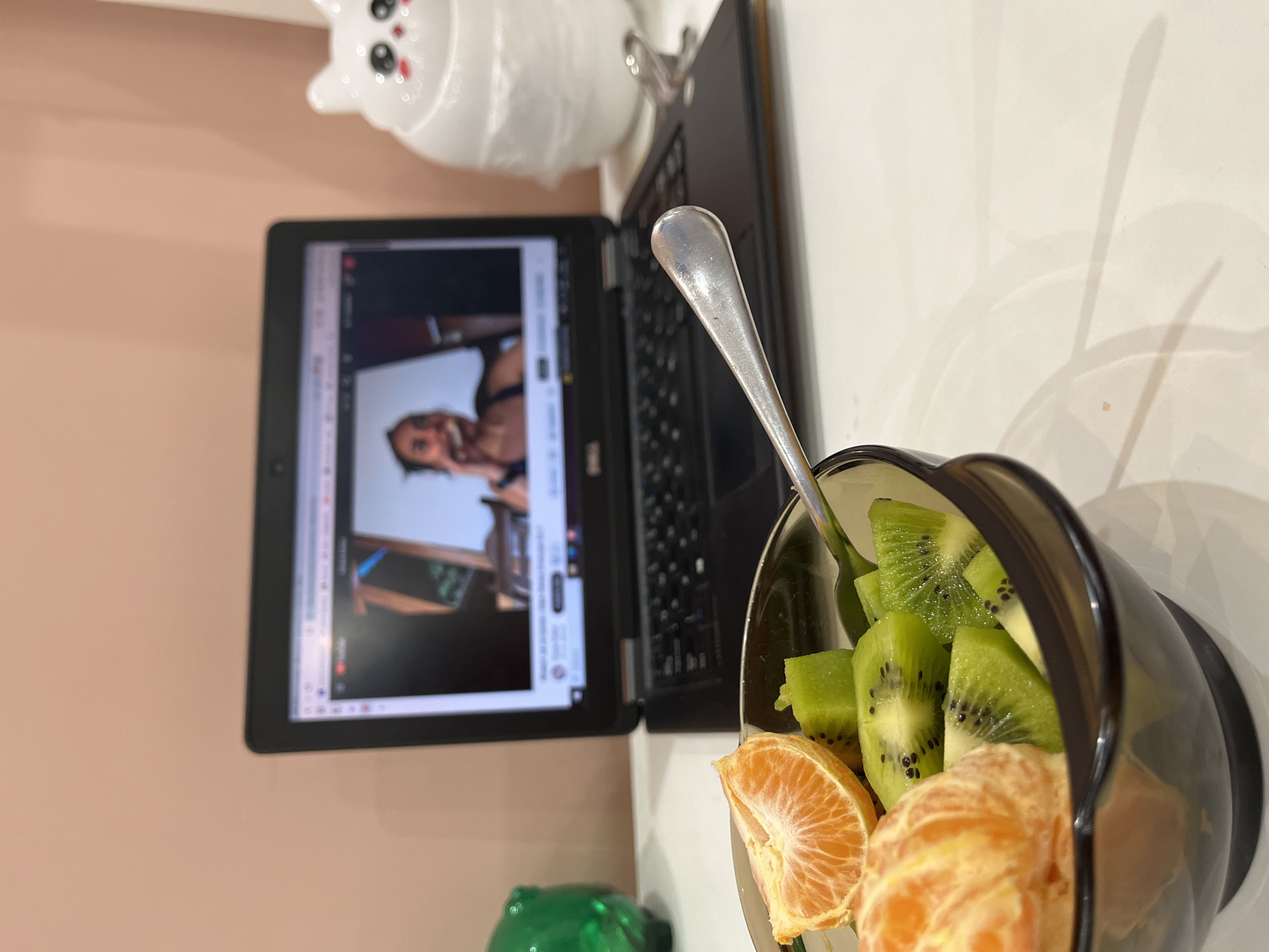 The image depicts an indoor scene in Pernik, Bulgaria, primarily focused on a tabletop setup. In the foreground, a translucent dark brown bowl contains a fruit salad composed of peeled mandarin orange segments and uniformly diced green kiwi fruit, with a silver spoon resting inside. Centered in the mid-ground is an open black Dell laptop, its screen displaying an active YouTube video. The video content shows a blurred, chest-up view of an individual, likely female, with dark hair, wearing a dark top, and appearing to hold a light-colored object near her mouth. To the right of the laptop, a white, stylized, glossy cat-shaped figurine, featuring black eyes and pink cheek accents, is visible, partially encased in clear plastic wrapping. A green, translucent object, possibly a container, is partially visible on the far left side of the background. The background wall is a smooth, light peach or pink color, and the surface beneath the objects is white.