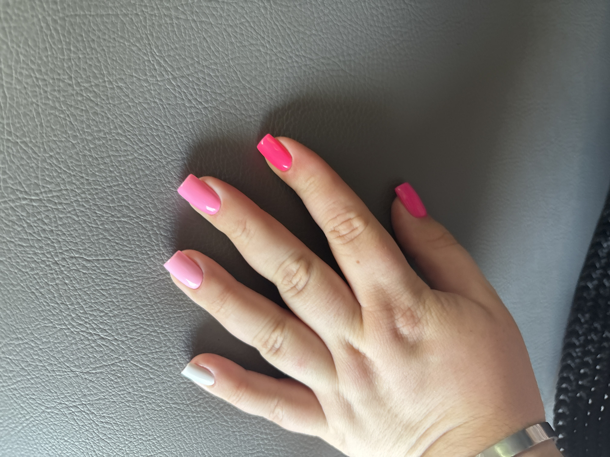 A person's hand, with long, square-shaped artificial nails painted in various bright colors, is resting on a textured gray surface, possibly a couch or car seat. The nails are painted in white, light pink, bright pink, and neon pink. The hand is positioned with the palm facing down, and the fingers are slightly spread. The background is out of focus, but appears to be a continuation of the gray textured surface. There is a black woven material visible at the bottom edge of the image, and a silver bracelet on the person's wrist. The image appears to be taken indoors in natural light. There is no specific information about the location in Мастоцкі сельскі Савет, Belarus.