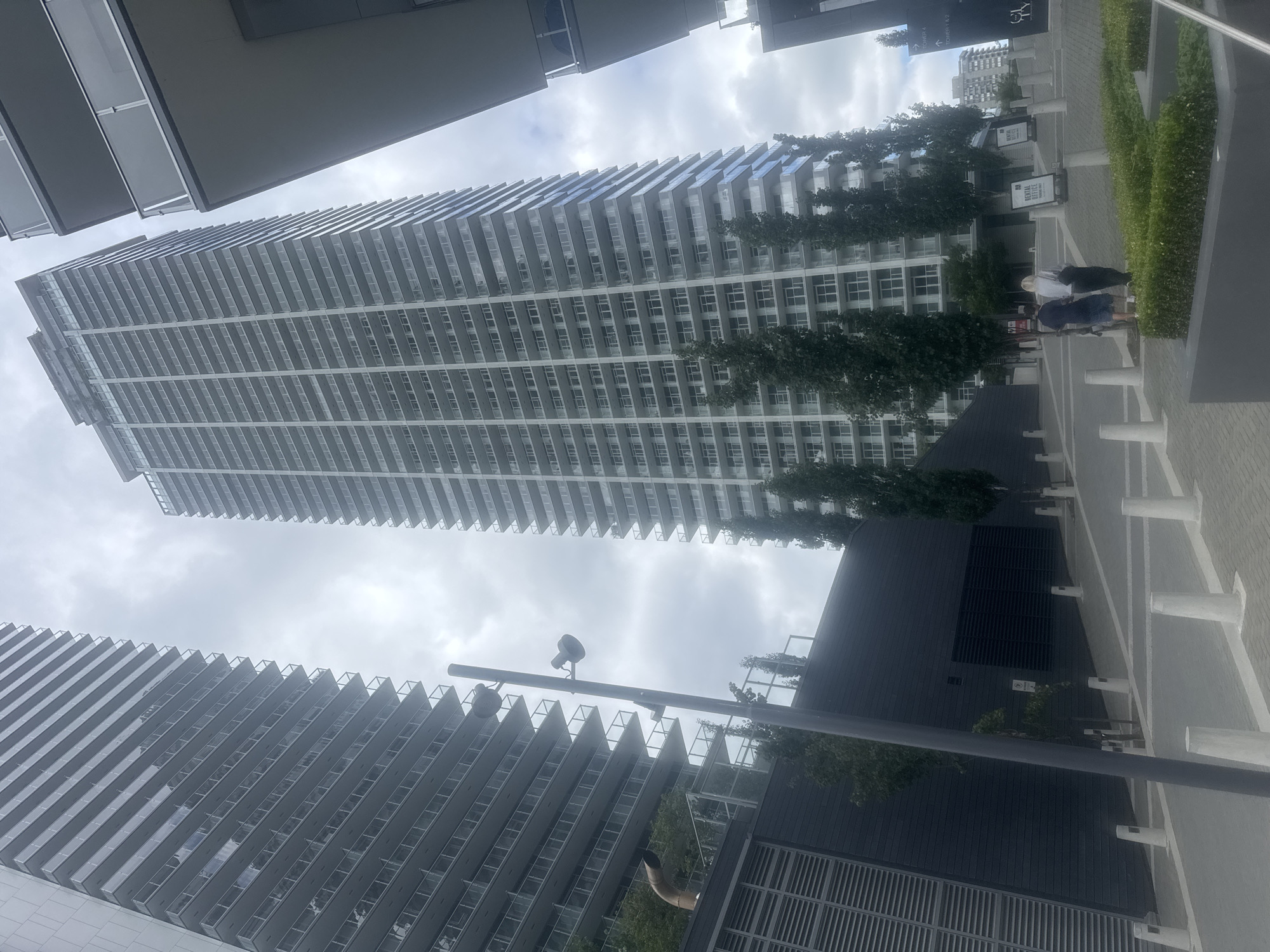 The image shows a street view in Burnaby, Canada, with two modern high-rise residential buildings dominating the frame. The building on the left is closer and has a facade of glass balconies and light-colored panels. The taller building in the center is also a high-rise with numerous balconies. In the foreground, a paved walkway is lined with bollards and a low hedge. Two individuals, a man and a woman, are walking away from the camera on the walkway. A street lamp with two fixtures is positioned to the left of the center. The environment appears to be an urban setting with a cloudy sky overhead.