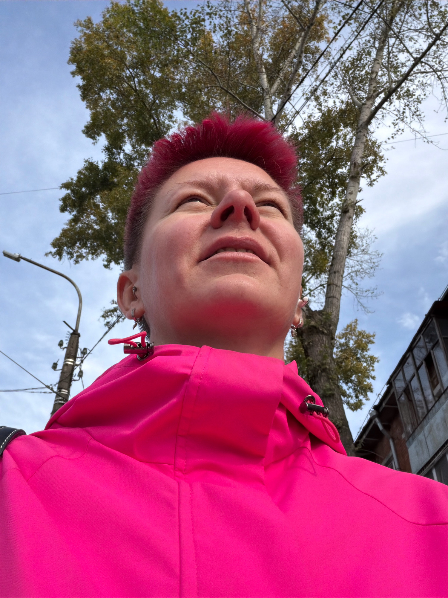 A low-angle, close-up outdoor shot shows a person with short, bright pink hair looking upwards, their face partially obscured by the collar of a vibrant pink jacket. The jacket has a metallic zipper pull on the shoulder. In the background, a tall tree with sparse autumn foliage stands against a cloudy sky. To the left, a street lamp on a concrete pole is visible. To the right, a portion of a weathered building with multiple windows is apparent. The scene appears to be on a street in the city of Chaikovsky, Russia.