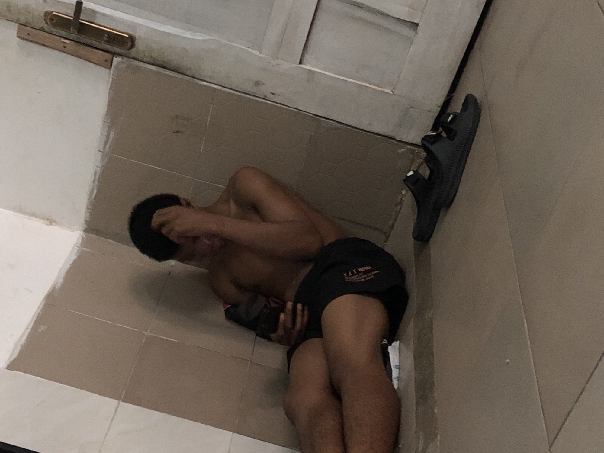 An indoor scene depicts a person, appearing male, lying on their side on a tiled floor. The individual is shirtless and wears dark shorts with white text visible on the left leg, which reads "XXX METAN..." and "TRAPPED...". Their head is partially obscured by an arm or hand, and they appear to be holding a dark-colored object, possibly a phone, with their other hand.
The floor consists of light brown or beige square tiles, and the walls are light-colored, possibly white or off-white, with some visible grime or marks, particularly near the floor line and a door frame. A brass-colored door handle or latch is prominently visible on a light-colored wooden door or door frame at the top of the image. Leaning against a wall or door frame to the right is a single black sandal or slide. The overall impression is a somewhat sparse or utilitarian room or hallway. Given the provided context, the location is Ado, Nigeria. The person's posture could suggest rest, sleep, or a state of discomfort.