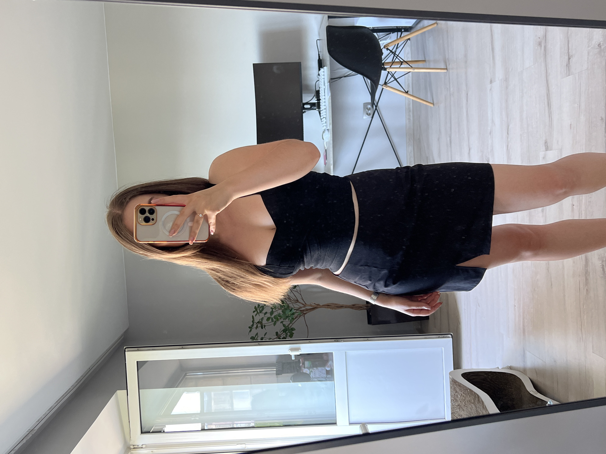 A woman is taking a mirror selfie in a room. She is wearing a black dress and holding a phone in her right hand to take the picture. Her left arm is resting on her head. The room appears to be a home office or living space. In the background, a desk with a computer monitor and keyboard is visible, along with a black chair with wooden legs. A window with blinds is also in the background, reflecting some light. A potted plant sits on a surface near the window. The flooring is light-colored wood. The time of day appears to be daytime, judging by the natural light coming from the window. There are no visible signs of text in the image. The location is noted as Ternopil, Ukraine.