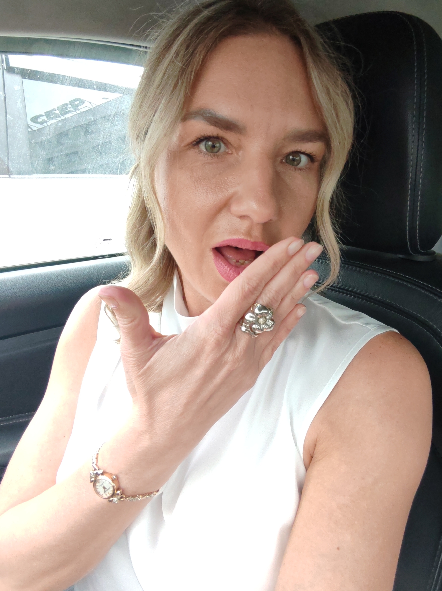 A woman with blonde hair is sitting in a car, looking at the camera. She is wearing a white sleeveless shirt and has her right hand raised to her mouth. She is wearing a large ring on her right index finger and a bracelet on her left wrist. The interior of the car, including a black leather headrest, is visible behind her. Outside the car window, a building with the word "SEEP" visible on it can be seen. The day appears to be overcast.