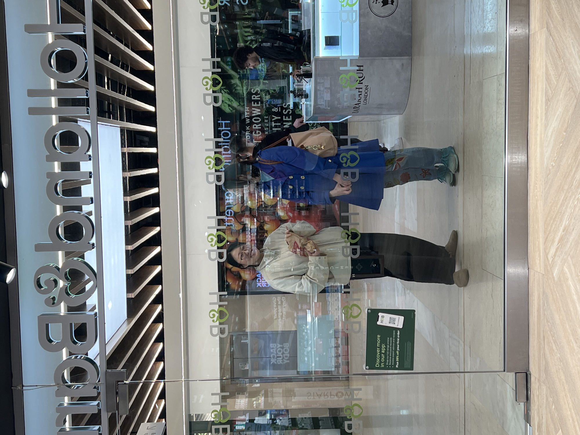 The image shows the storefront of a Holland & Barrett (H&B) store, recognizable by its signage. The store appears to be located in a shopping center in Epsom and Ewell, United Kingdom. The store's glass doors reflect the street scene, obscuring the view inside. Two women are reflected in the glass doors. One is wearing a blue coat and floral jeans and carrying a tan purse, and the other is wearing a light-colored shirt. A man is standing inside the store in front of a counter. The store displays advertising for the Holland and Barrett app, which offers shopping and exclusive rewards. The interior of the store is only partially visible, with glimpses of shelves and products.