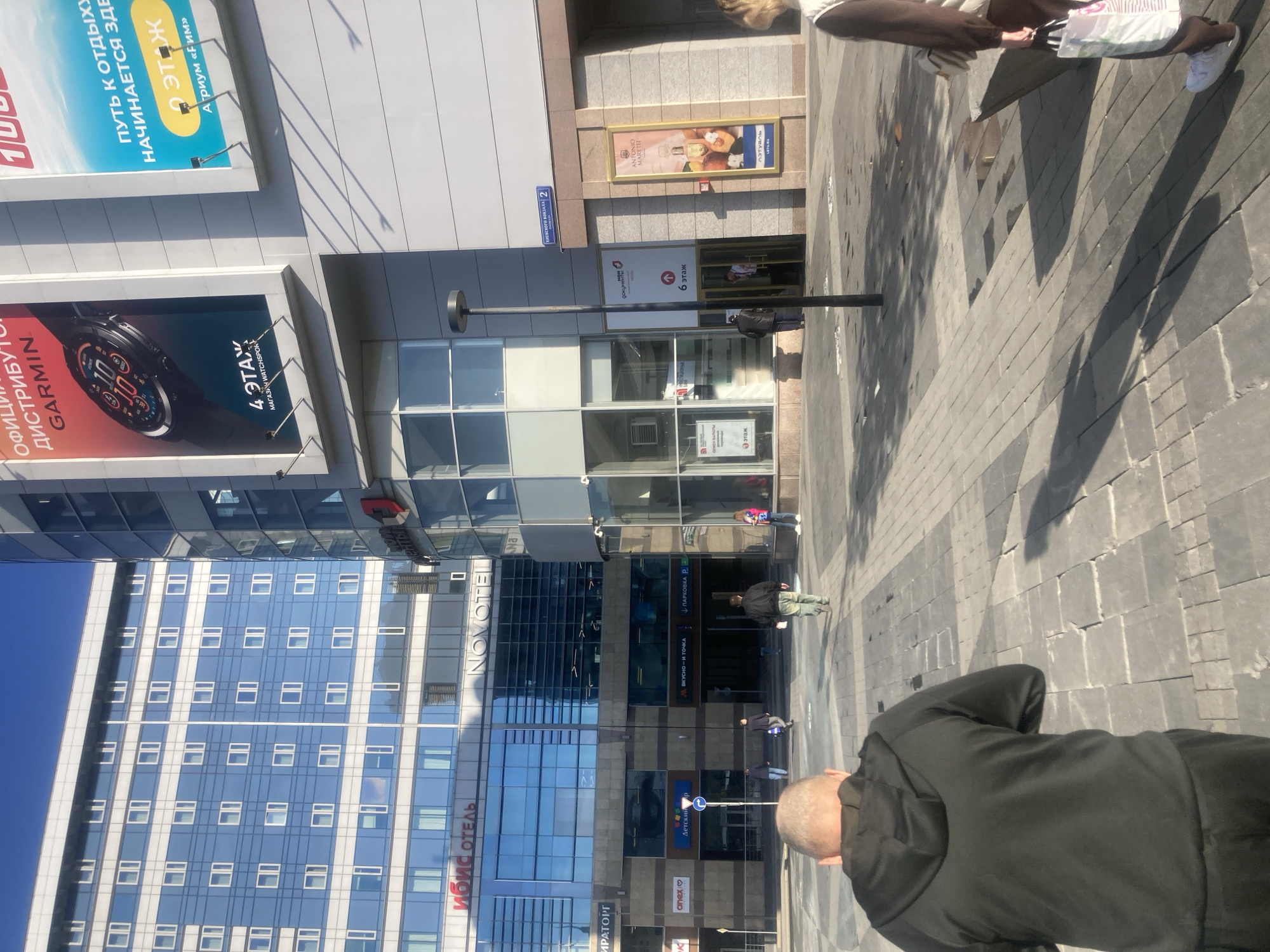 The image shows a street scene in Moscow, Russia, with a mix of modern buildings, advertisements, and pedestrians. The prominent architectural features include the ibis hotel and the Novotel hotel, which are multi-story buildings with reflective windows. There are also advertisements for Garmin watches, perfumes, and the fast-food chain "Vkusno i tochka" (formerly McDonald's). Street signs point to various locations such as "Children's" and "Parking". Several pedestrians are visible on the paved walkway, and one person is wearing a black jacket. The scene appears to be taken during the daytime under bright sunlight.