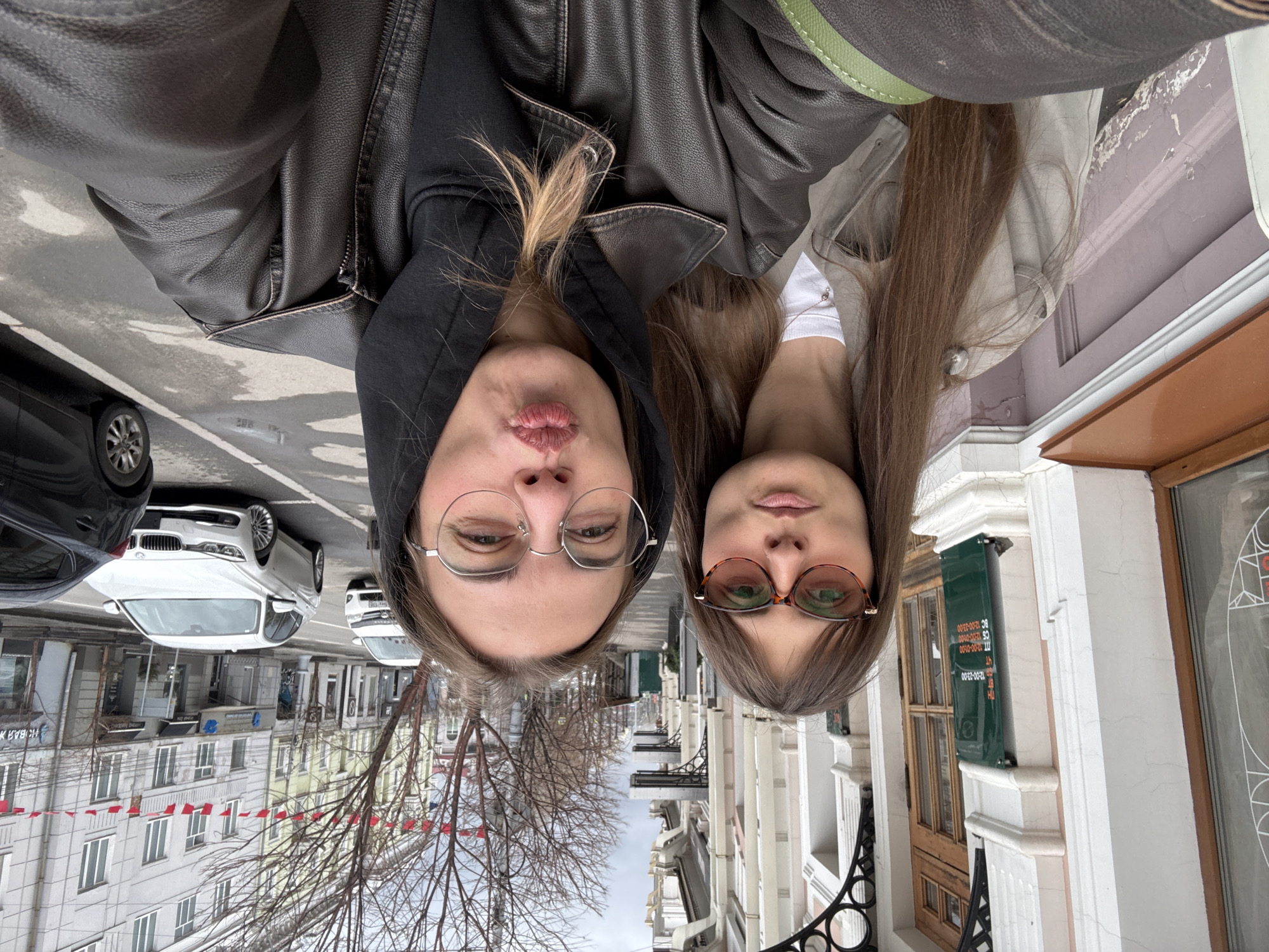 Two women are posing closely together for a selfie outdoors on a street. The woman on the left is wearing a black hooded jacket and round wire-rimmed glasses, puckering her lips. The woman on the right has straight brown hair and is wearing brown-rimmed glasses. Both are wearing leather jackets.
The background shows a street in Irkutsk, Russia. Cars are parked on the left side of the street. Buildings line both sides, with bare trees visible in the center. A street sign with flags is visible on the left. A green sign is on the right, and a wrought-iron fence is below the sign.
