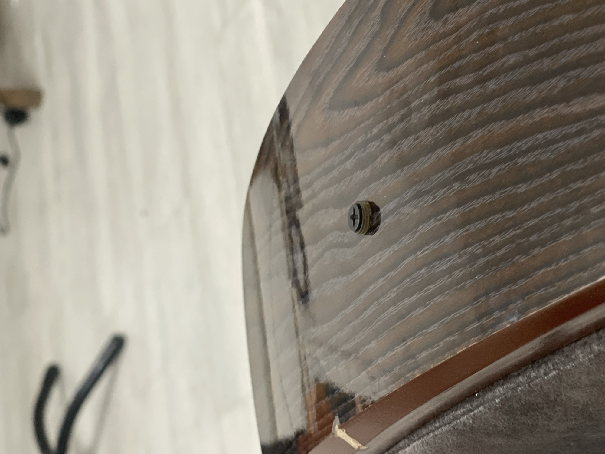The image shows a close-up of a dark brown, glossy wooden surface with a visible wood grain pattern. A single black screw with a cross-shaped head is partially embedded in the surface. The screw head is slightly raised and appears to be made of metal, possibly with some rust or grime on its edges. The wooden surface has a reflective sheen, indicating it might be a piece of furniture or a finished wood panel. In the background, a blurred black object, possibly a chair leg or a similar structure, is visible, along with a light-colored floor and a light wooden object. The overall scene appears to be indoors, in a well-lit environment. No specific location within Bishkek City, Kyrgyzstan can be determined from the visual information provided in the image.