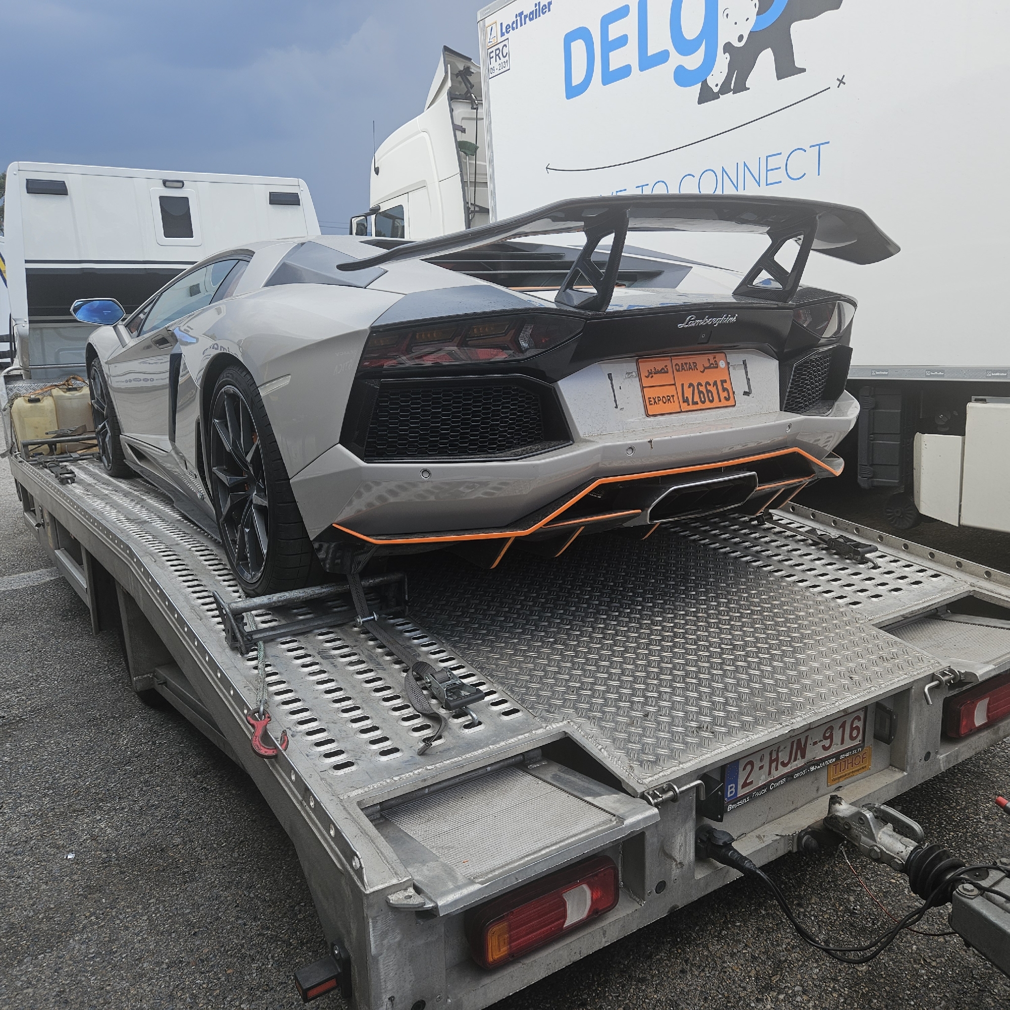 In the city of La Jonquera, Spain, a silver Lamborghini Aventador sports car is parked on a silver car carrier trailer. The car faces away from the camera, revealing its rear. It has a large black rear wing and orange accents on the diffuser and side skirts. The license plate on the rear of the car reads "EXPORT 426615" with Arabic text above it and "QATAR" visible. The trailer is connected to a white truck, part of which is visible in the background. The truck has blue lettering on its side that reads "DELG[image of a bear]Y". The trailer is equipped with straps to secure the car. The ground is asphalt. The sky is overcast.