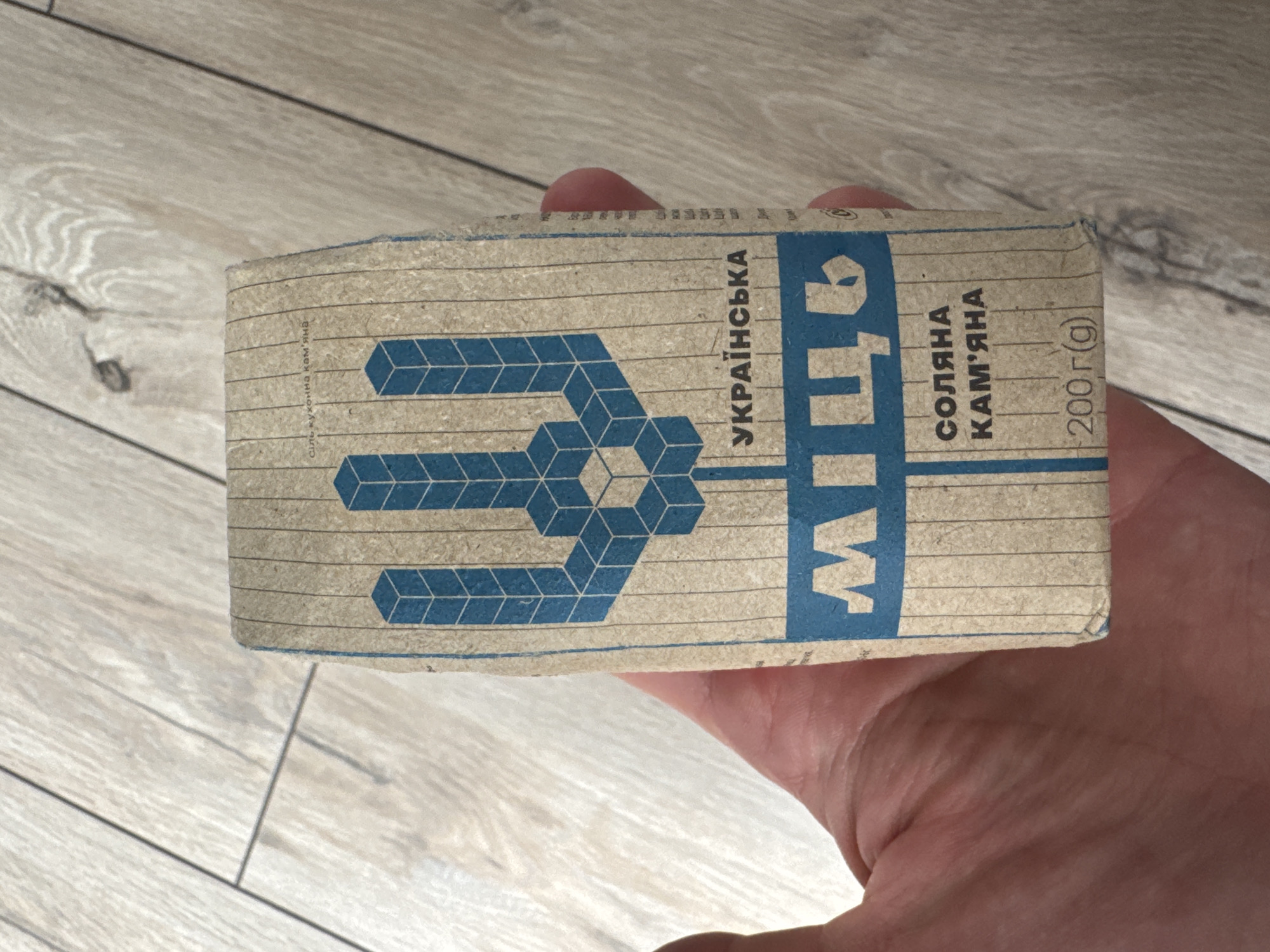 A hand holds a rectangular package of Ukrainian salt with blue printing on a brown background. The package is labeled "УКРАЇНСЬКА СІЛЬ СОЛЯНА КАМ'ЯНА 200 г (g)". The packaging features a blue trident design. The background is a light-colored wooden surface, possibly a floor. The location is unknown.