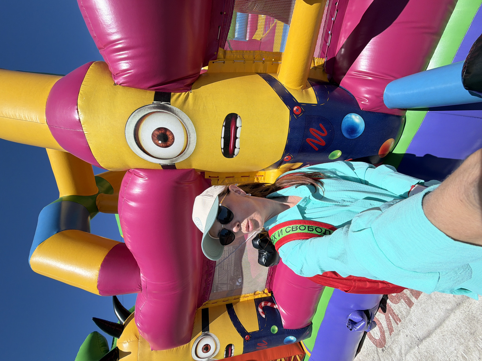A woman is taking a selfie in front of an inflatable Minion-themed bouncy castle. She is wearing a turquoise shirt, a beige baseball cap, and sunglasses, with a red backpack strapped on. The bouncy castle is colorful, with large depictions of Minions and other characters. The background is a clear blue sky. The scene appears to be outdoors on a sunny day.