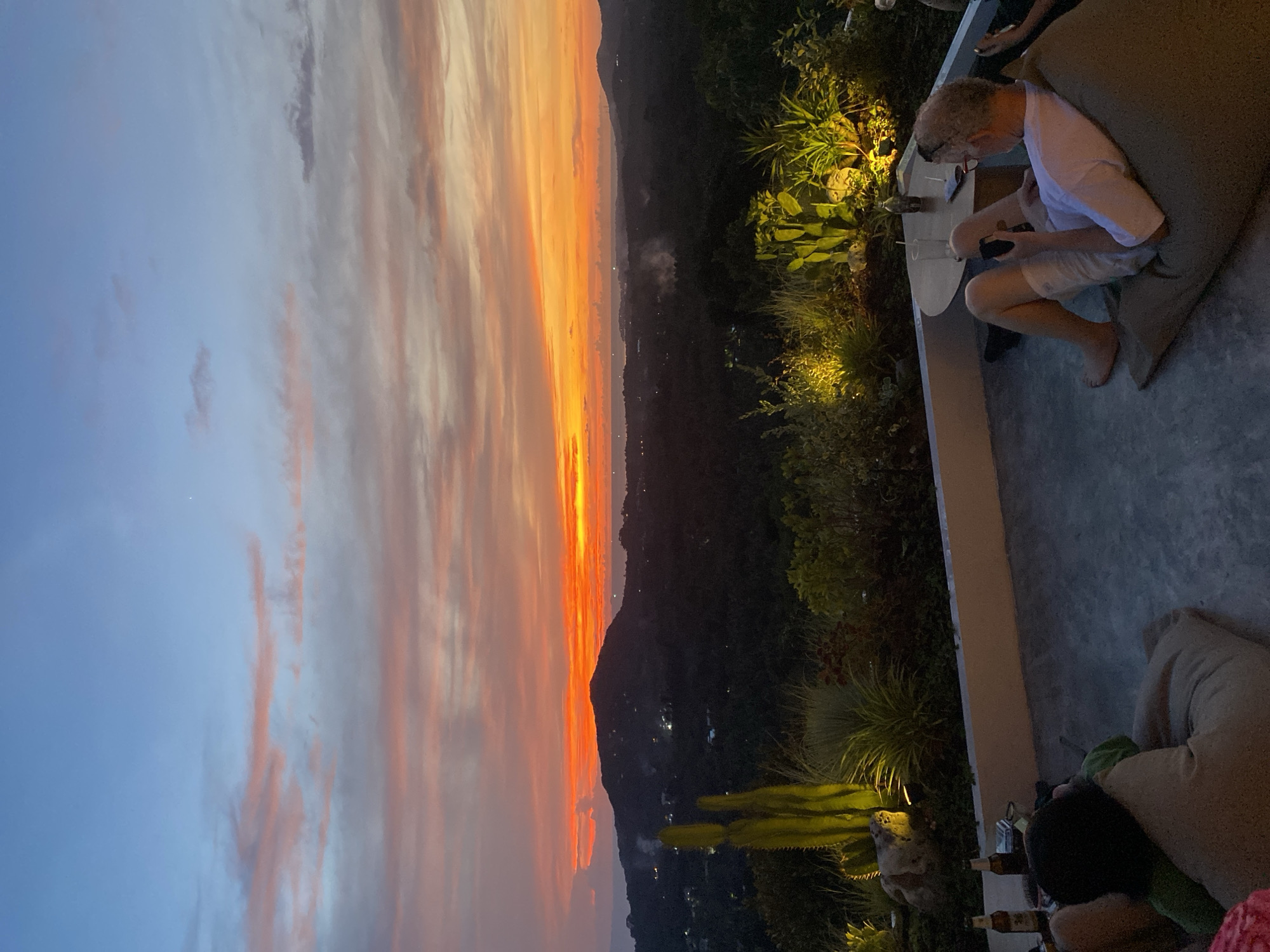 A vertical outdoor image shows a sunset over the hills of Baan Nai Suan, Thailand. The sky on the left shows hues of blue and pink. The setting sun casts a vibrant orange and red glow over the hills in the center of the image. The hills have sporadic patches of light suggesting the presence of small towns.
On the right side of the image, a terrace area is visible. A man with gray hair wearing a white shirt and light-colored shorts is seen seated on a brown beanbag chair. He is looking down and appears to be using a phone or small device. There is a table next to him with a few glasses on it. There are plants with upward-directed lighting along the terrace. Further down the terrace, at the bottom of the image, are beer bottles. A person with black hair is seated near the beer bottles.
