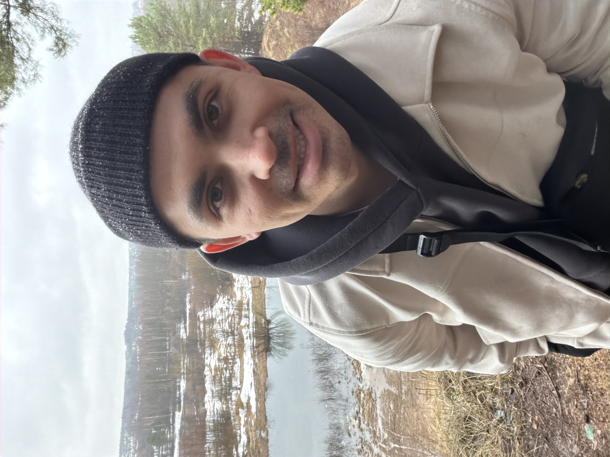 A man in a dark beanie, a black hooded sweatshirt, and a tan jacket is taking a selfie. The background shows a gray sky, trees, and a body of water with patches of snow on the banks. Based on visual cues (e.g., architecture and landscape), the location may be Izhevsk, Russia.