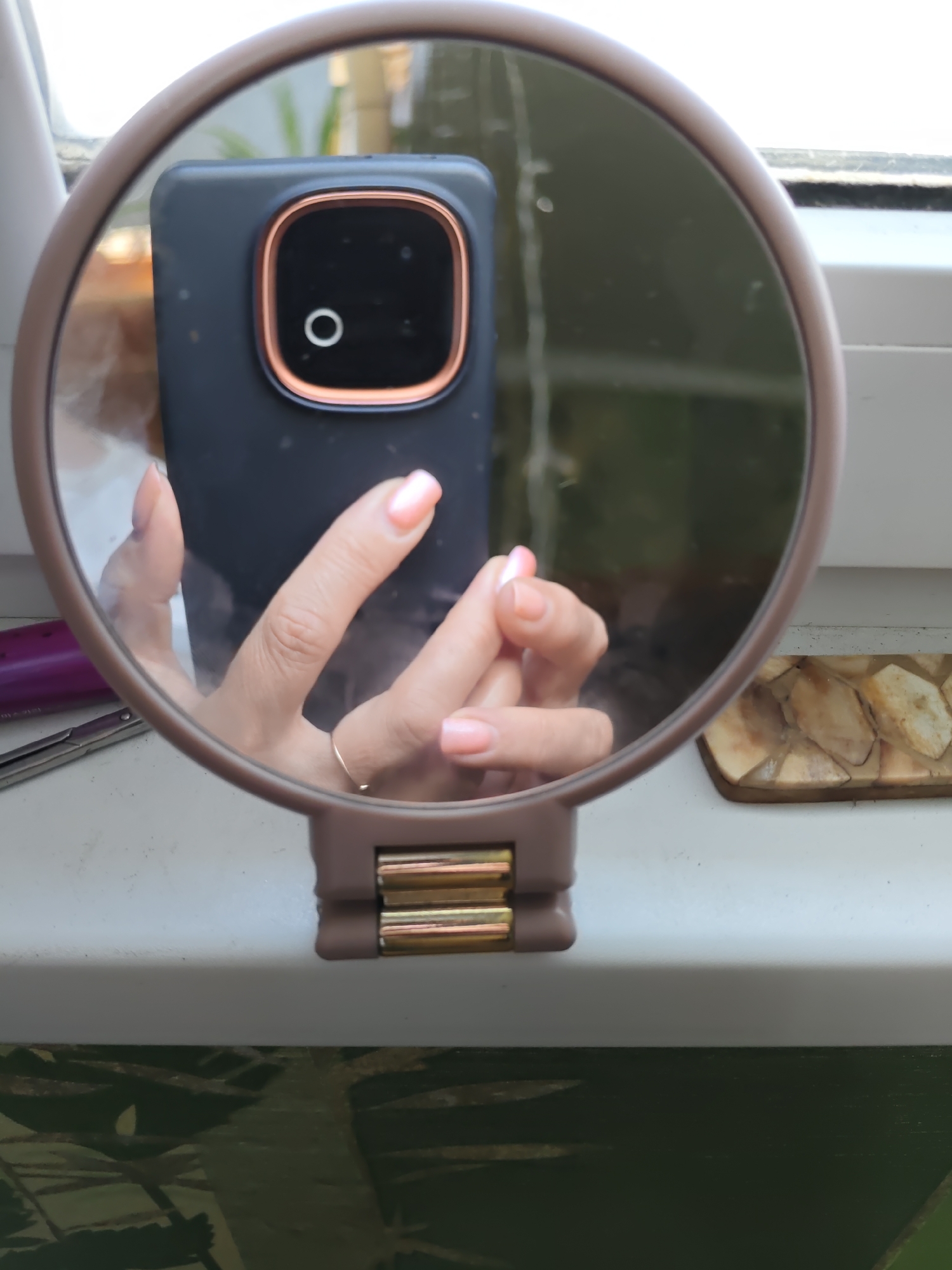 A reflection in a hand-held mirror shows a person's left hand, which has light pink manicured nails, holding a dark smartphone with a copper-colored camera housing. The mirror itself is round with a light brown frame and a visible hinge mechanism on its base. To the left of the mirror, a pair of scissors and a purple object are partially visible. To the right of the mirror, a decorative rectangular object made of light-colored, irregular pieces is placed. The mirror is positioned on a light-colored surface, possibly a windowsill, which overlooks a blurred green background. The overall scene is indoors, likely in a domestic setting. The city of Rostov-on-Don, Russia, is not explicitly visible or implied in this image.