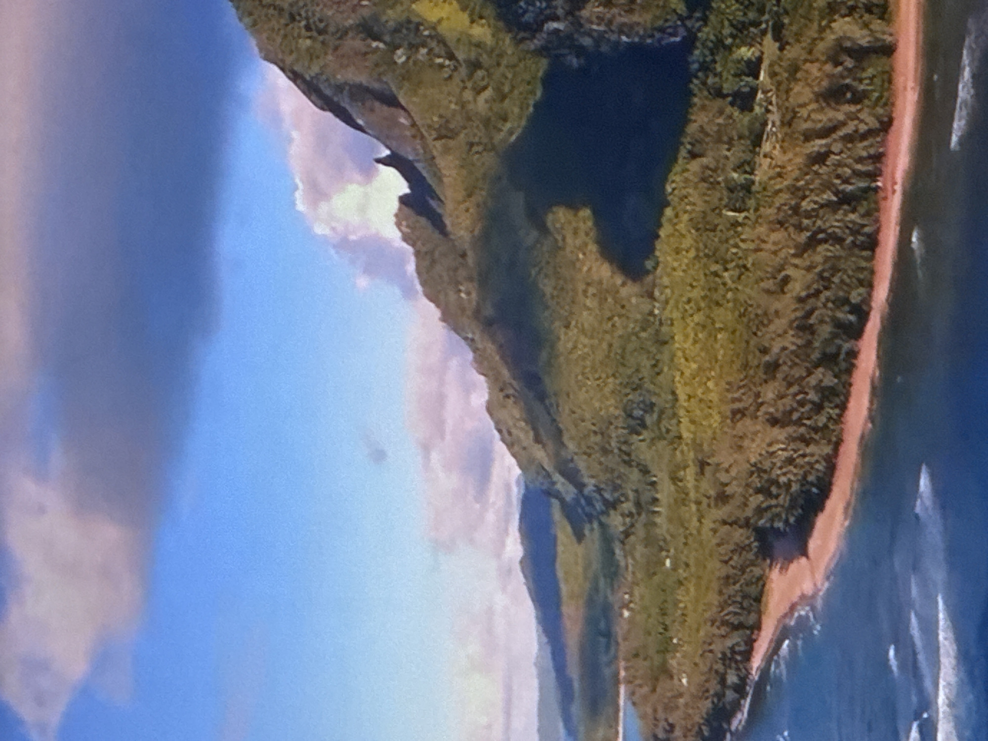 An aerial view captures a dramatic coastline with lush, green mountains meeting a sandy beach and the blue ocean. The sky above is a mix of bright blue and dark, brooding clouds, with streaks of pink and white suggesting the presence of sunlight breaking through. A prominent shadow is cast across the lower portion of the mountains, indicating the time of day and the position of the sun. The water shows gentle waves lapping at the shore, and a stretch of reddish-pink sand forms a narrow strip between the vegetation and the sea. The landscape appears natural and undeveloped, with no visible signs of human settlement or activity in the immediate vicinity.