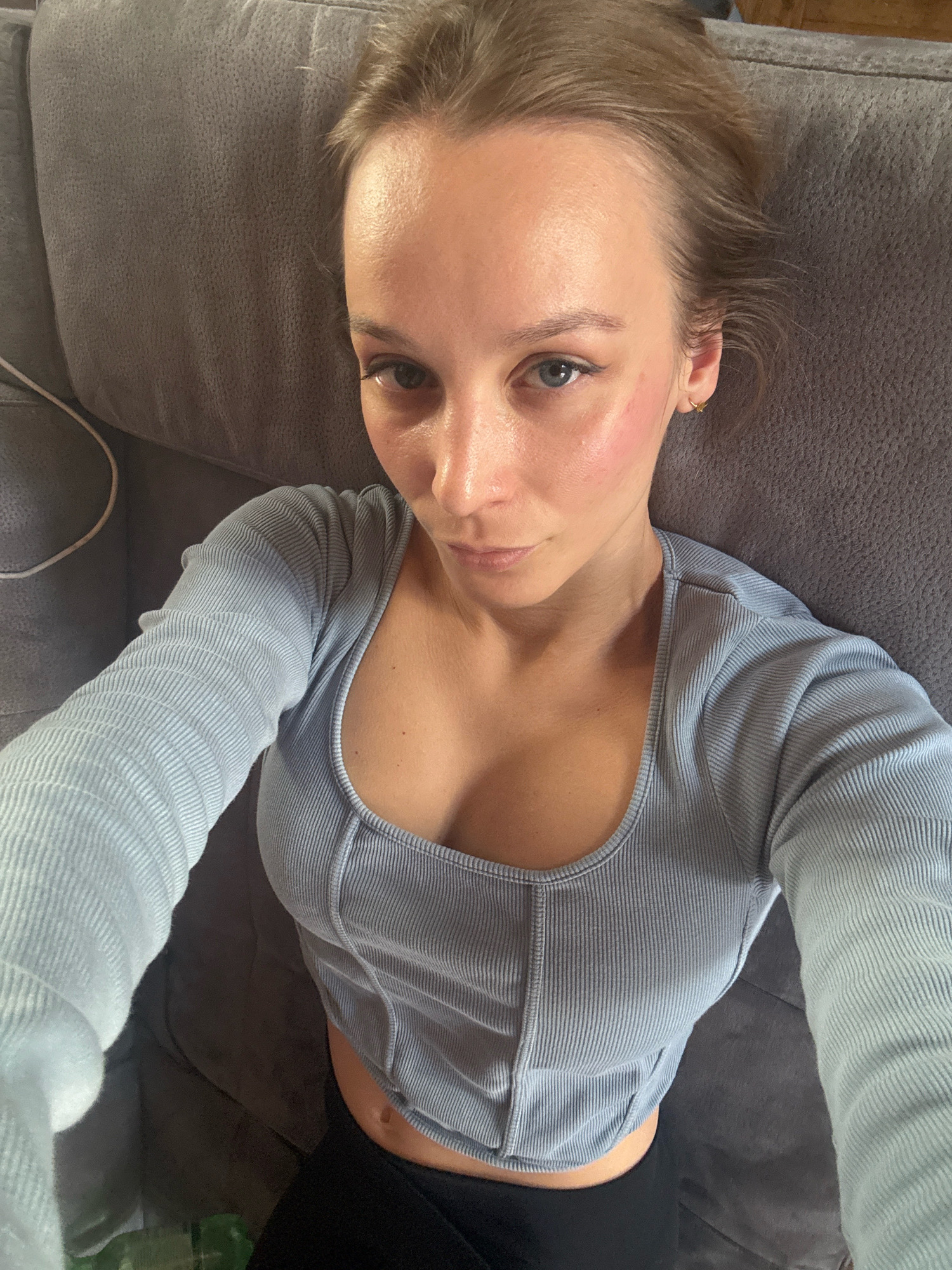 A young woman with blonde hair pulled back is lying on a grey couch, taking a selfie. She is wearing a light blue ribbed long-sleeved top and black leggings. Her head is tilted towards the camera, and she has blue eyes and rosy cheeks. The lighting suggests an indoor setting, possibly a living room. The scene is devoid of any identifiable landmarks of Überherrn, Germany.